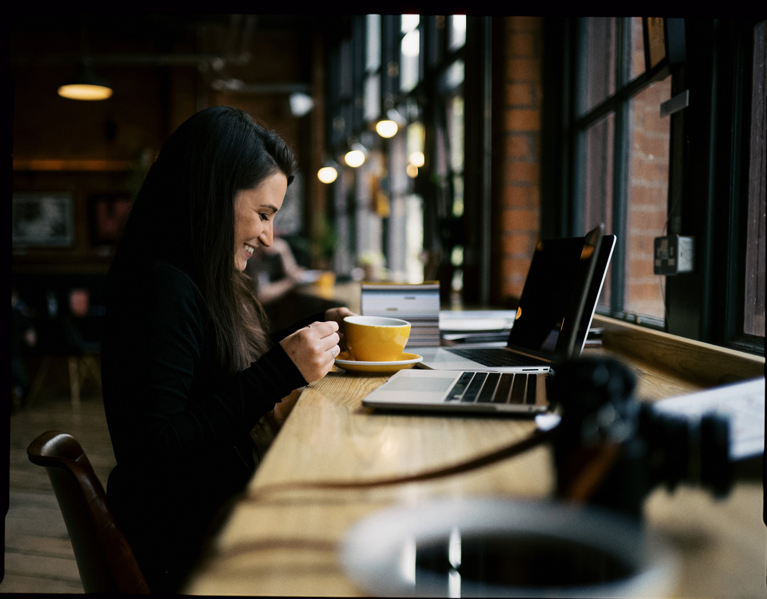 A woman with long dark hair laughing while holding a yellow coffee mug in a coffee shop or cafe with large windows, a laptop, and a stack of books on a wooden table.