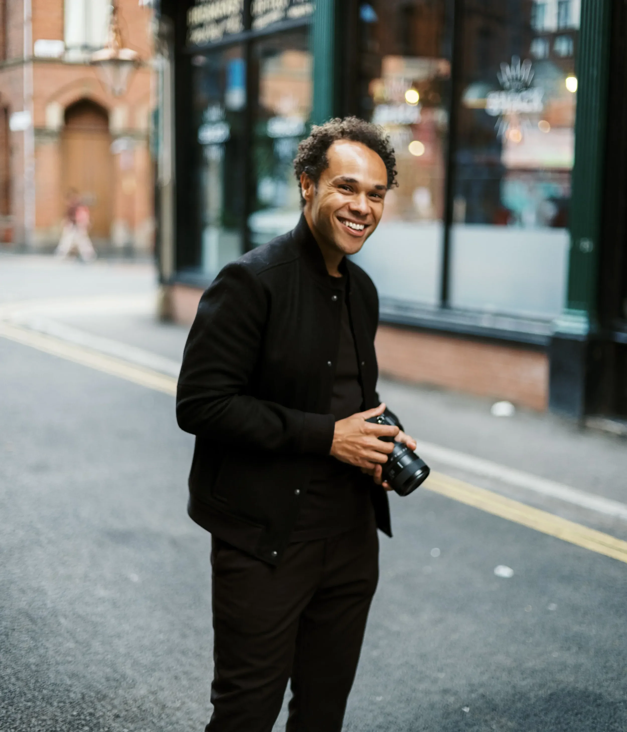 A man smiling on a city street holding a camera.