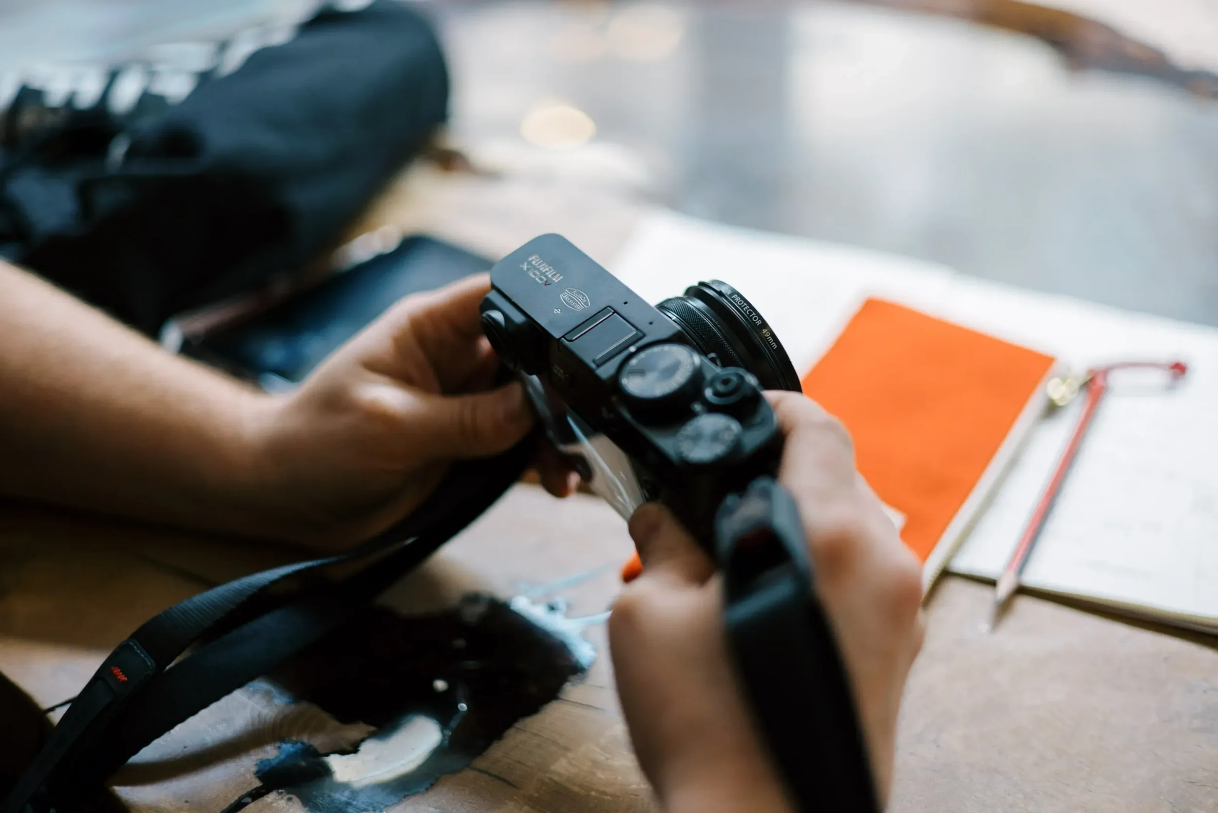 Person holding a camera over a wooden table with an open notebook and a pen, with a bag and a phone in the background.
