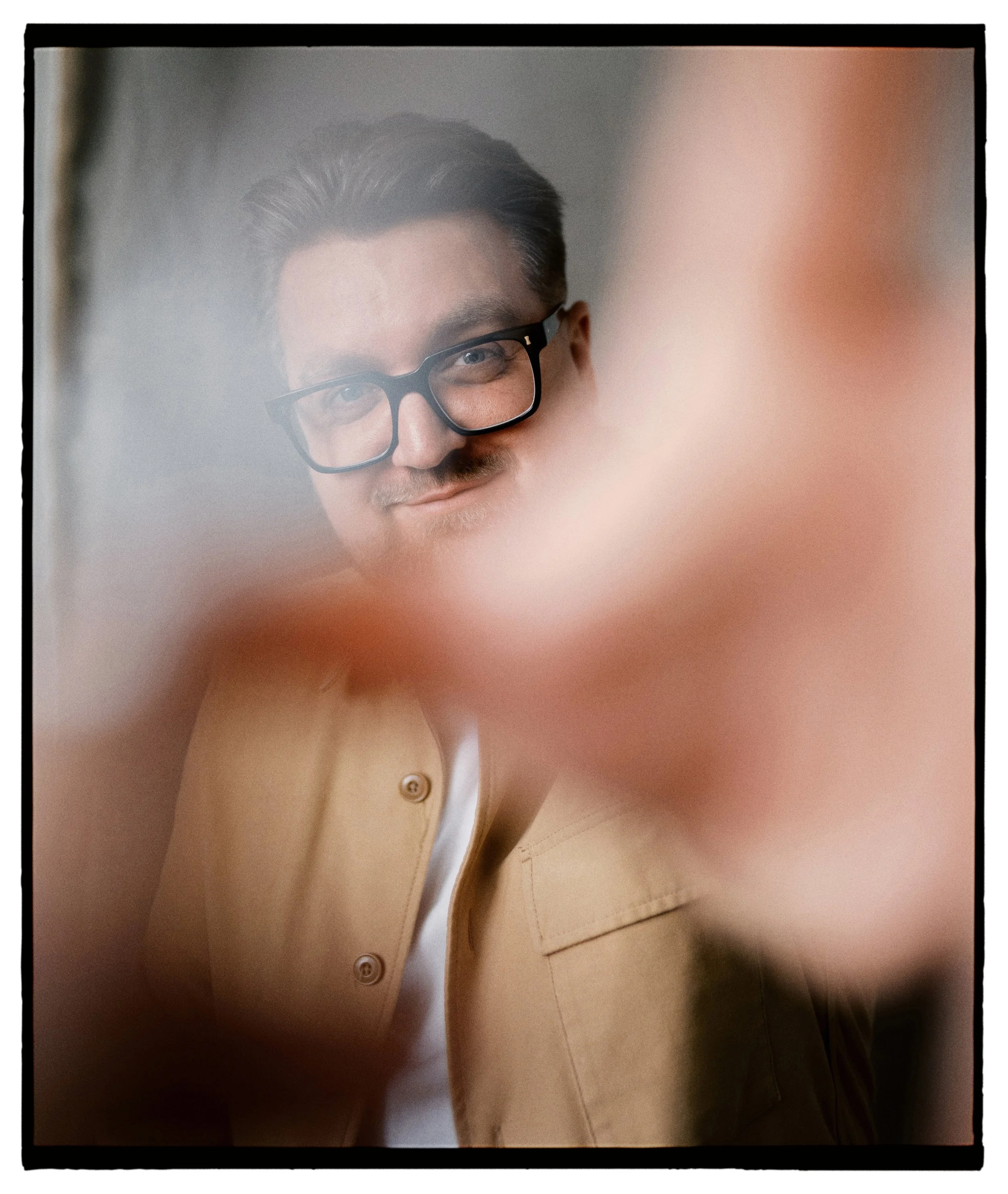 A man with glasses smiling, looking at the camera through a blurred hand in the foreground.