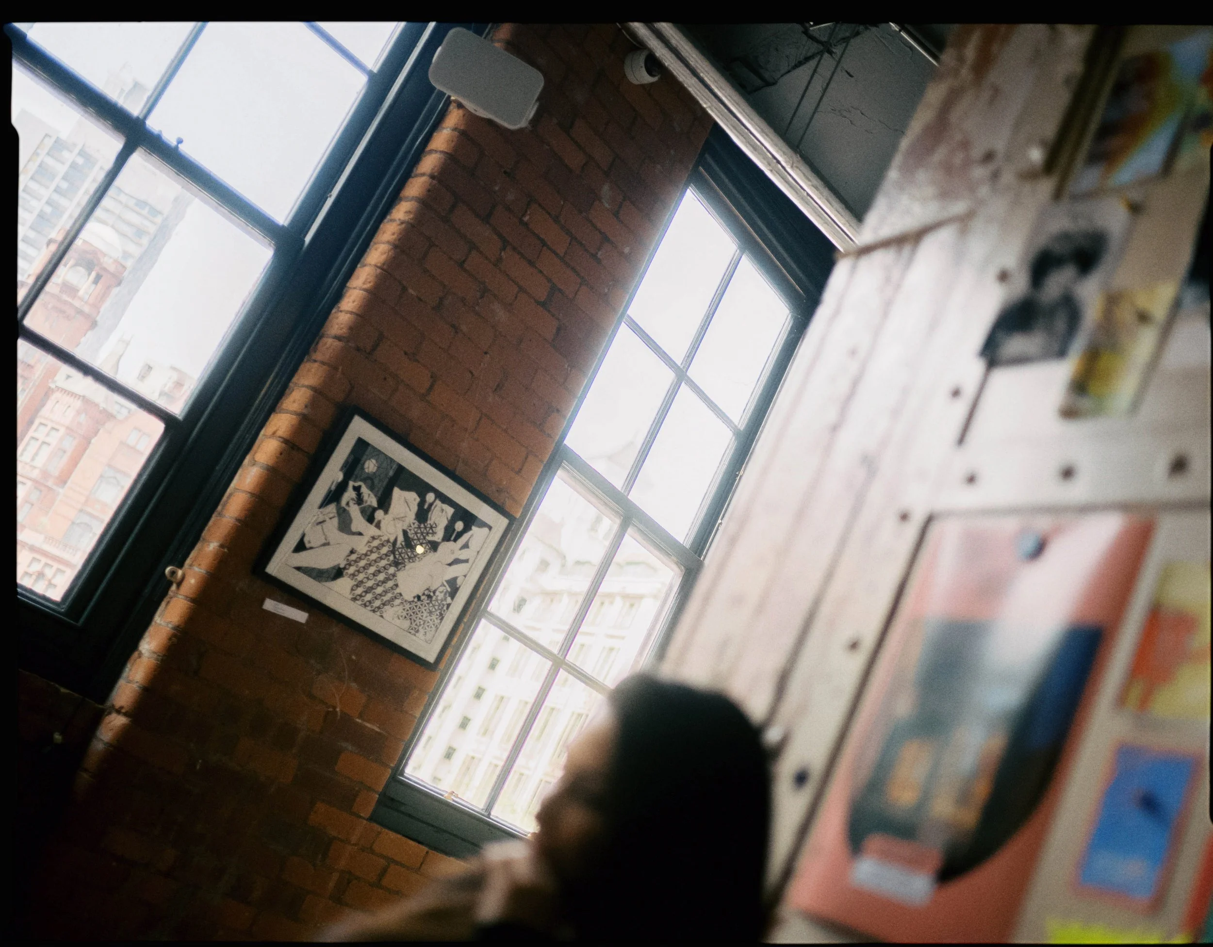 Interior of a room with brick walls and large windows showing a city view. A framed black-and-white artwork hangs on the wall. Part of a person's head is visible at the bottom of the image, and a wooden board with various items is also in the scene.