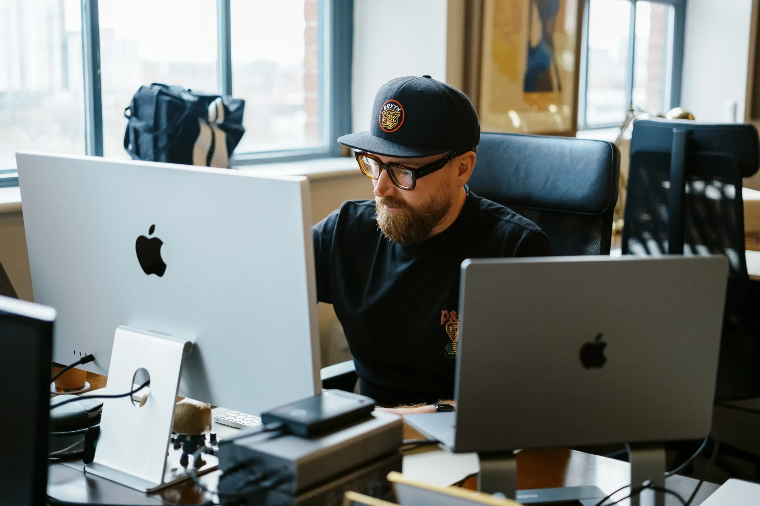 A man with a beard, glasses, wearing a black cap and black t-shirt, working at a desk with multiple Apple monitors in a modern office with large windows.