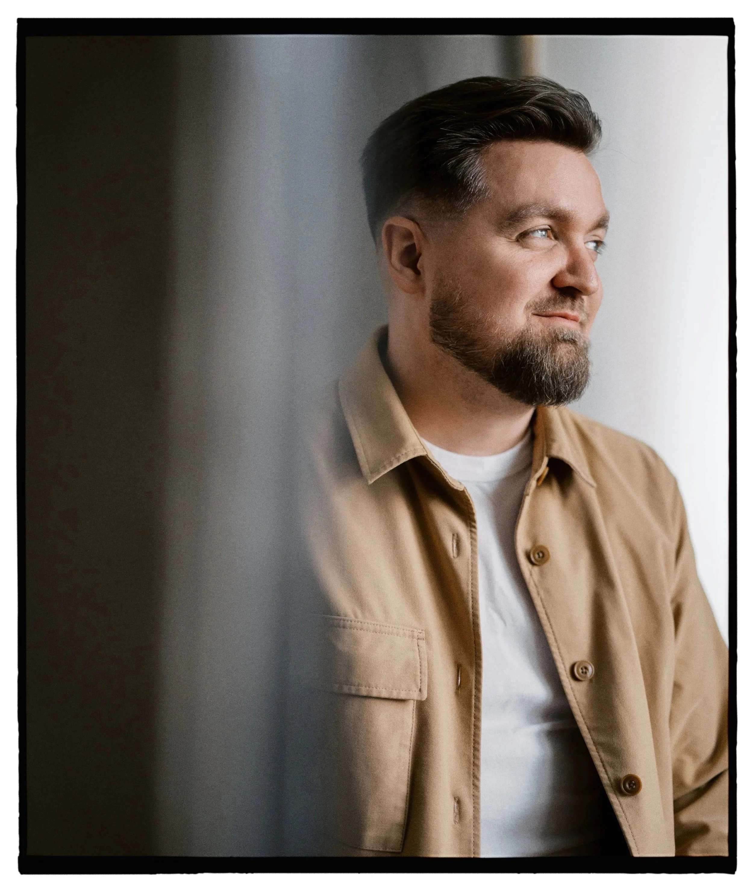 A man with a beard and styled hair looking out the window, wearing a beige jacket over a white shirt.