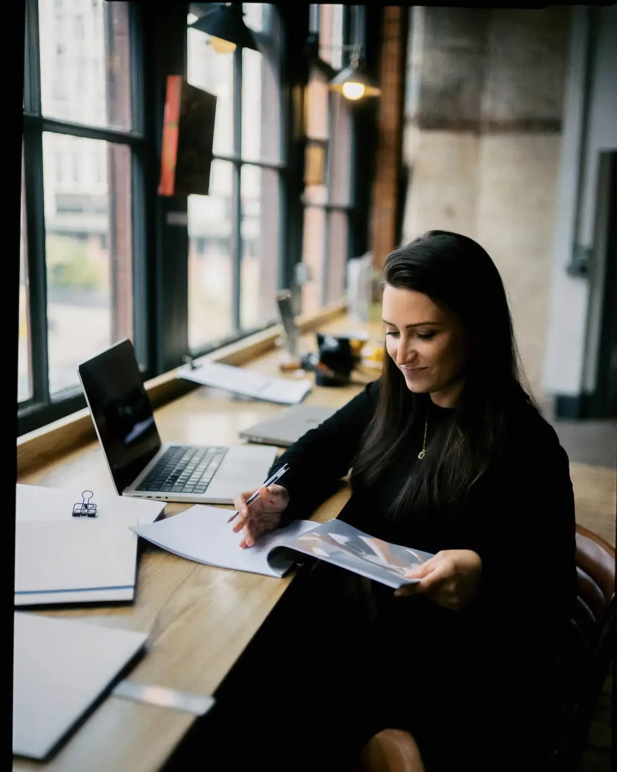 A woman sitting at a wooden desk in a brightly lit room, working with papers and a pen, with a laptop open beside her and documents spread out on the desk.