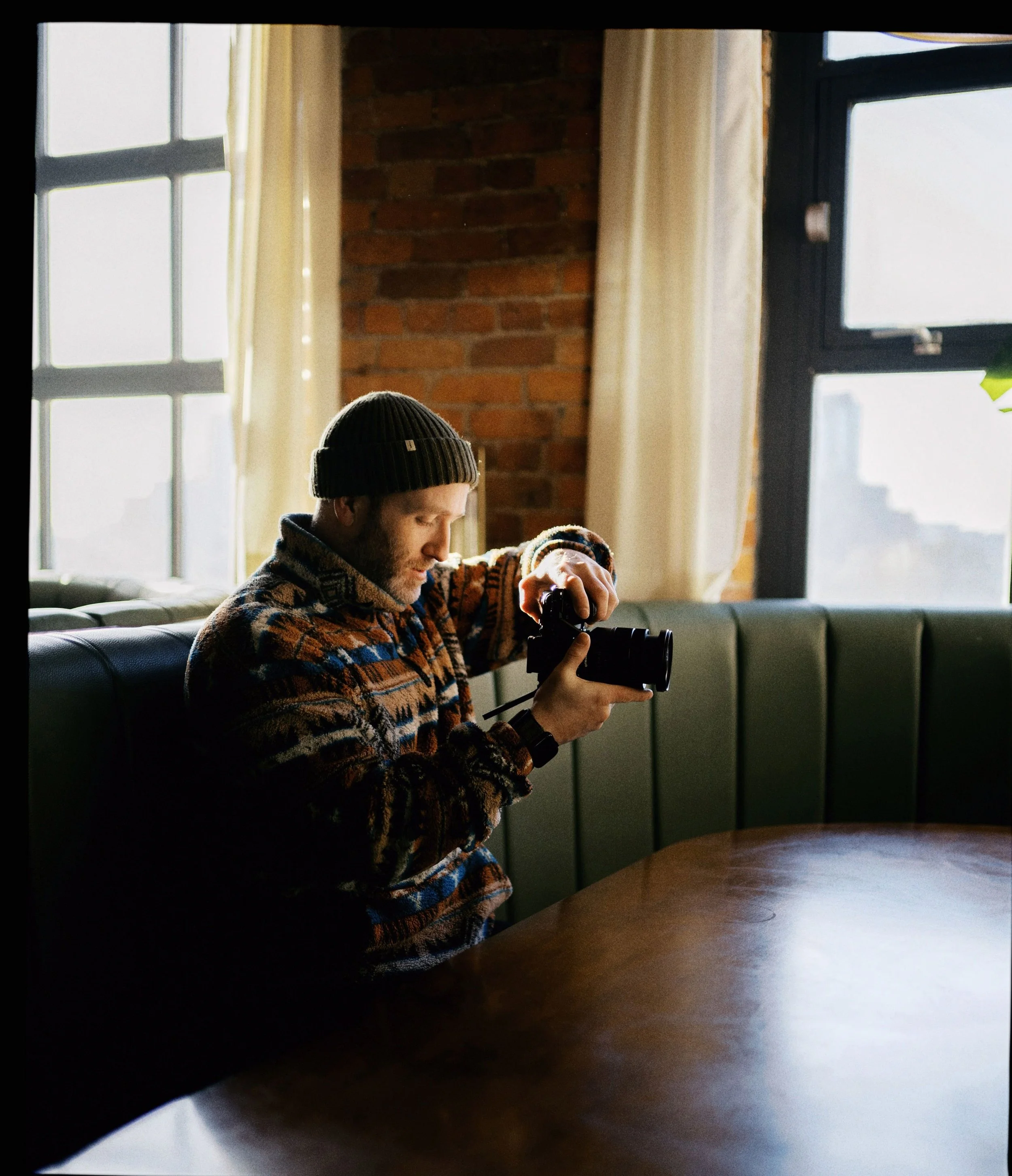 Man wearing a winter hat and patterned sweater taking a photo with a camera indoors near large windows and a brick wall.