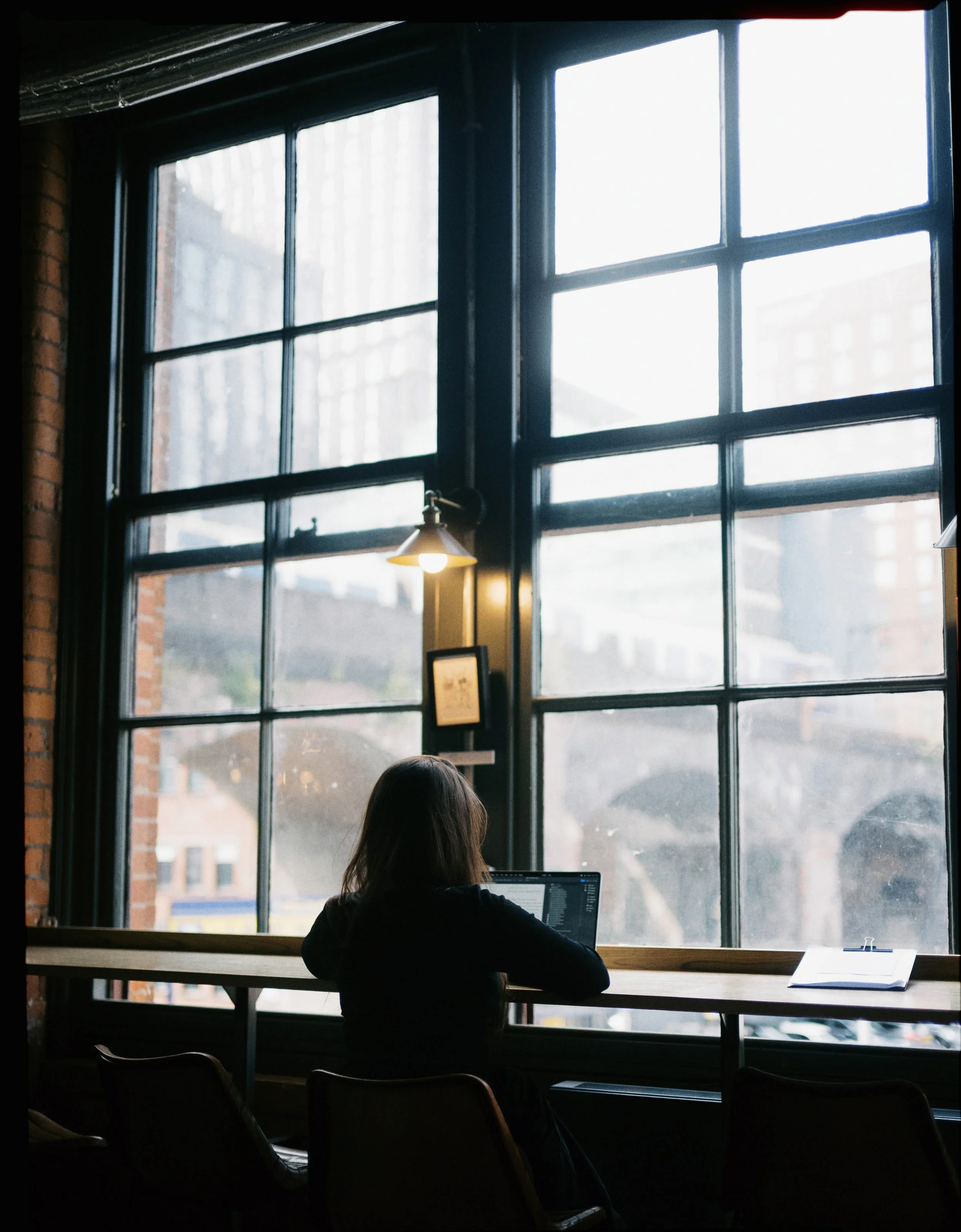 A woman sitting at a high wooden table near large industrial-style windows in a dimly lit cafe, working on a laptop.