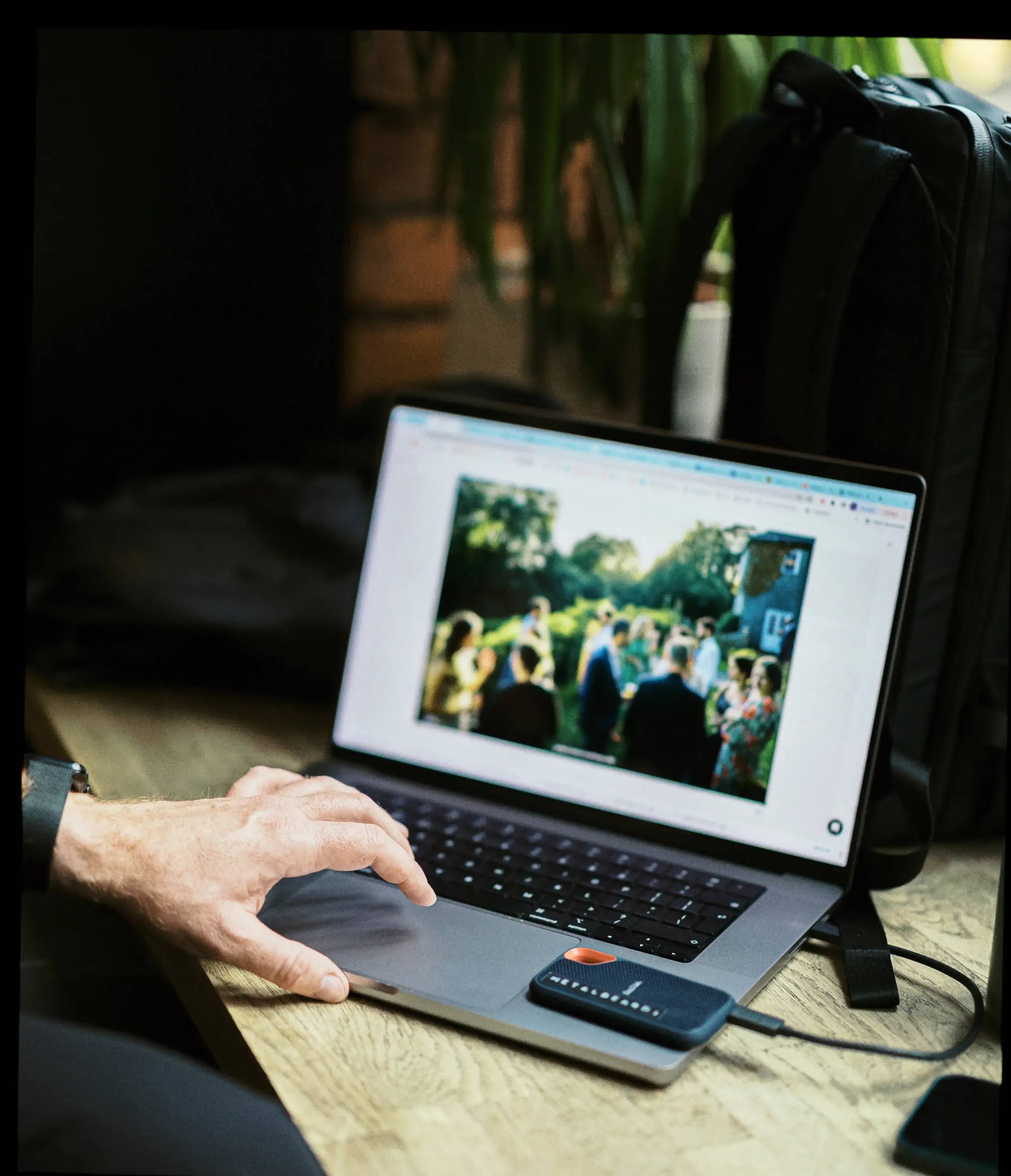 A person working on a laptop, viewing an outdoor event with people gathered and trees in the background.