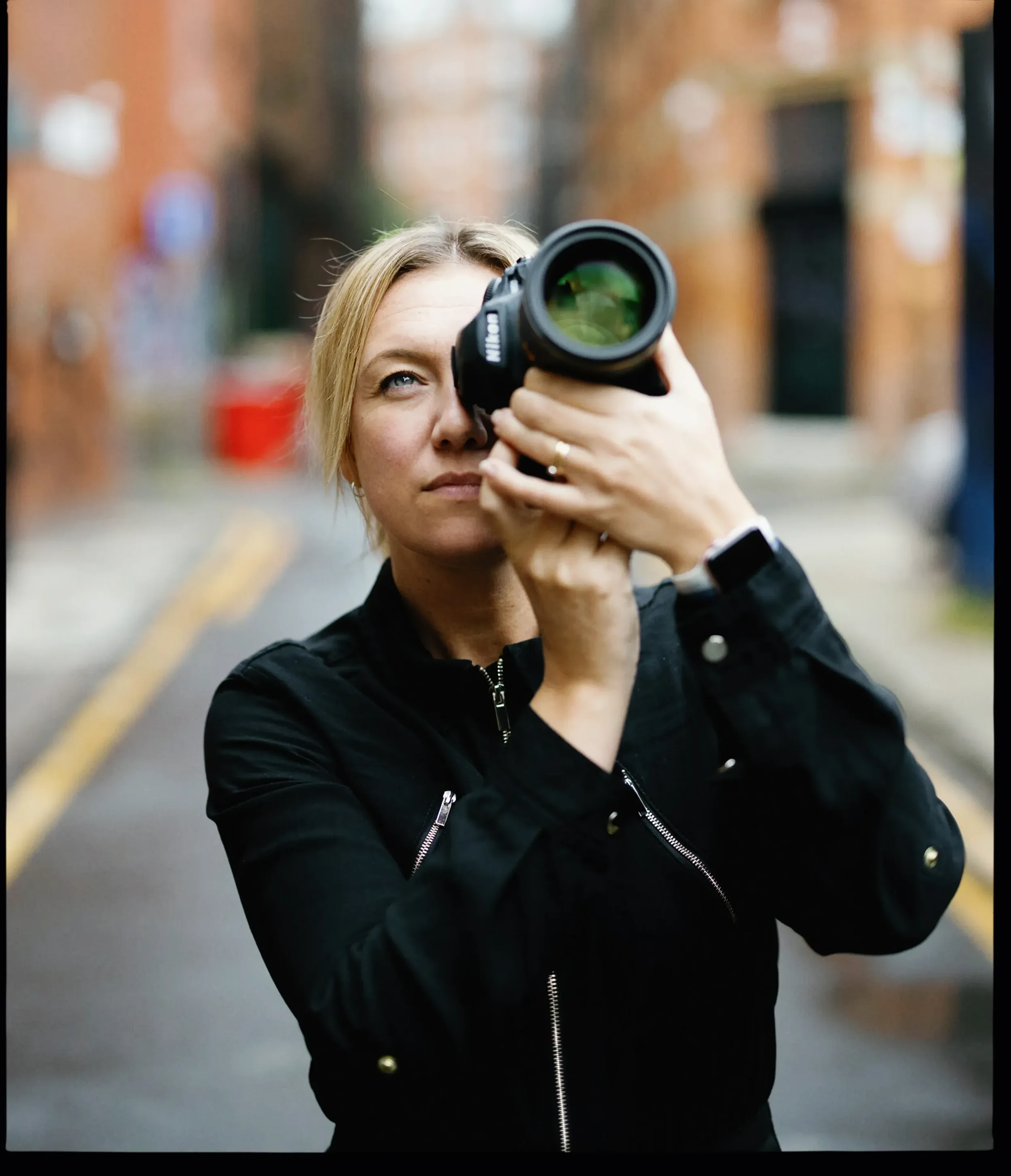 A woman with blonde hair takes a photo with a camera in an alleyway with brick buildings.