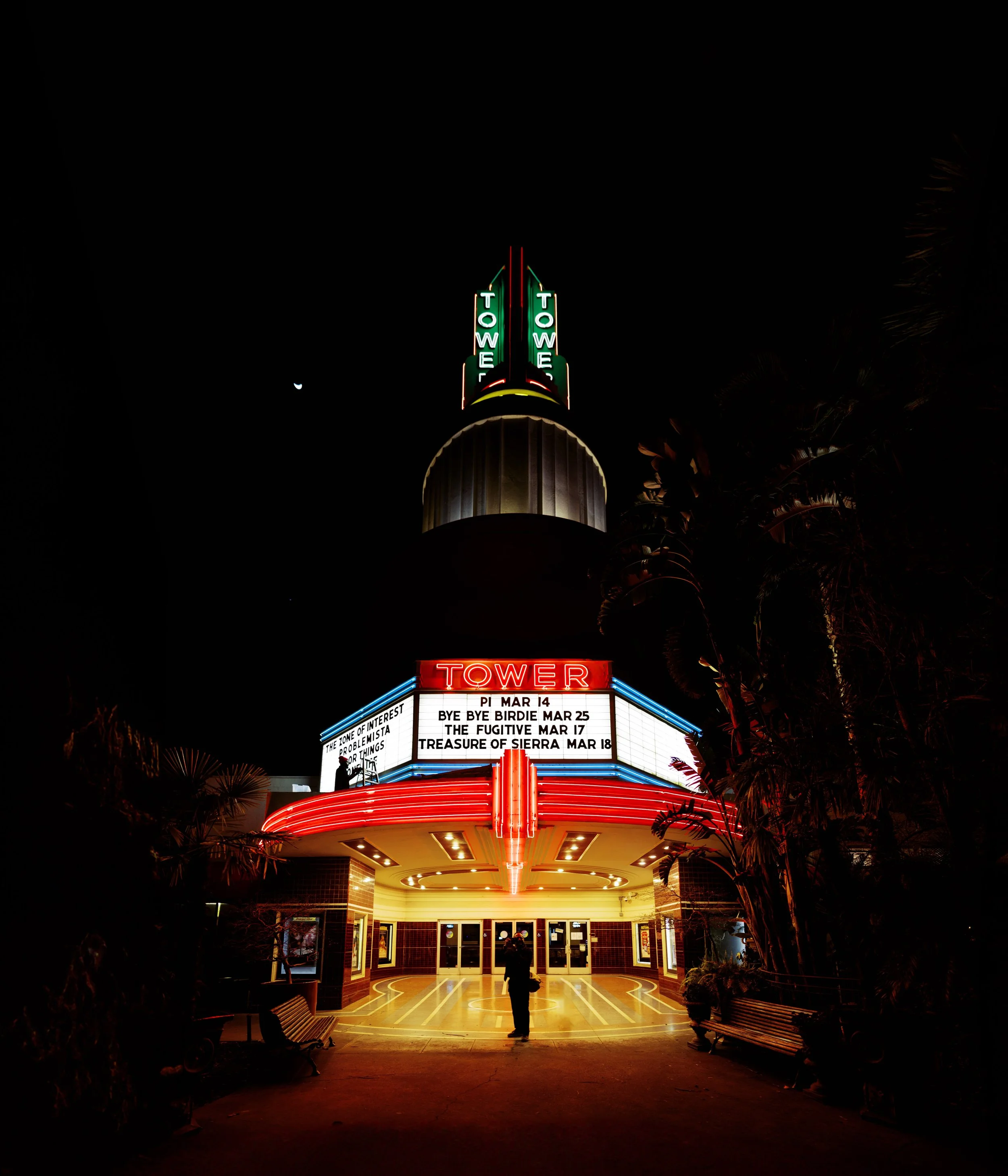 Night view of the Tower Theater with illuminated marquee, neon lights, and a person standing in front of the entrance.