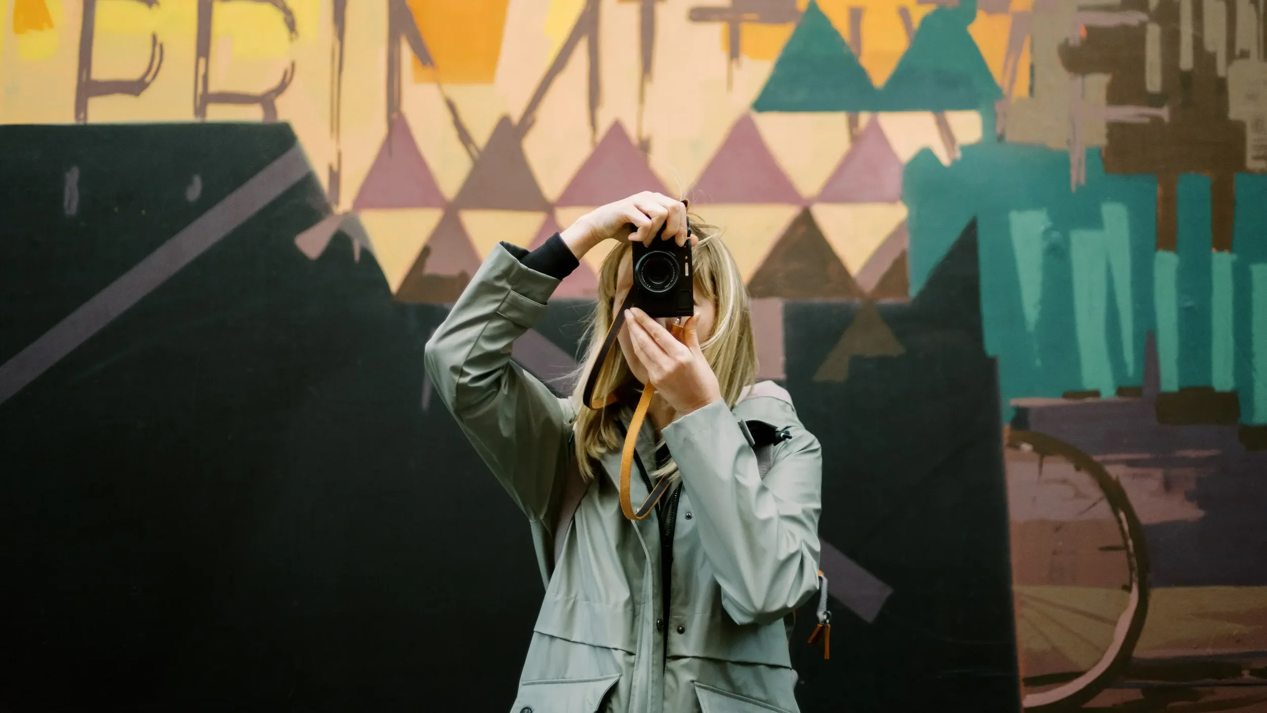 Woman with blonde hair, wearing a gray jacket, taking a photo with a camera in front of a colorful geometric mural.