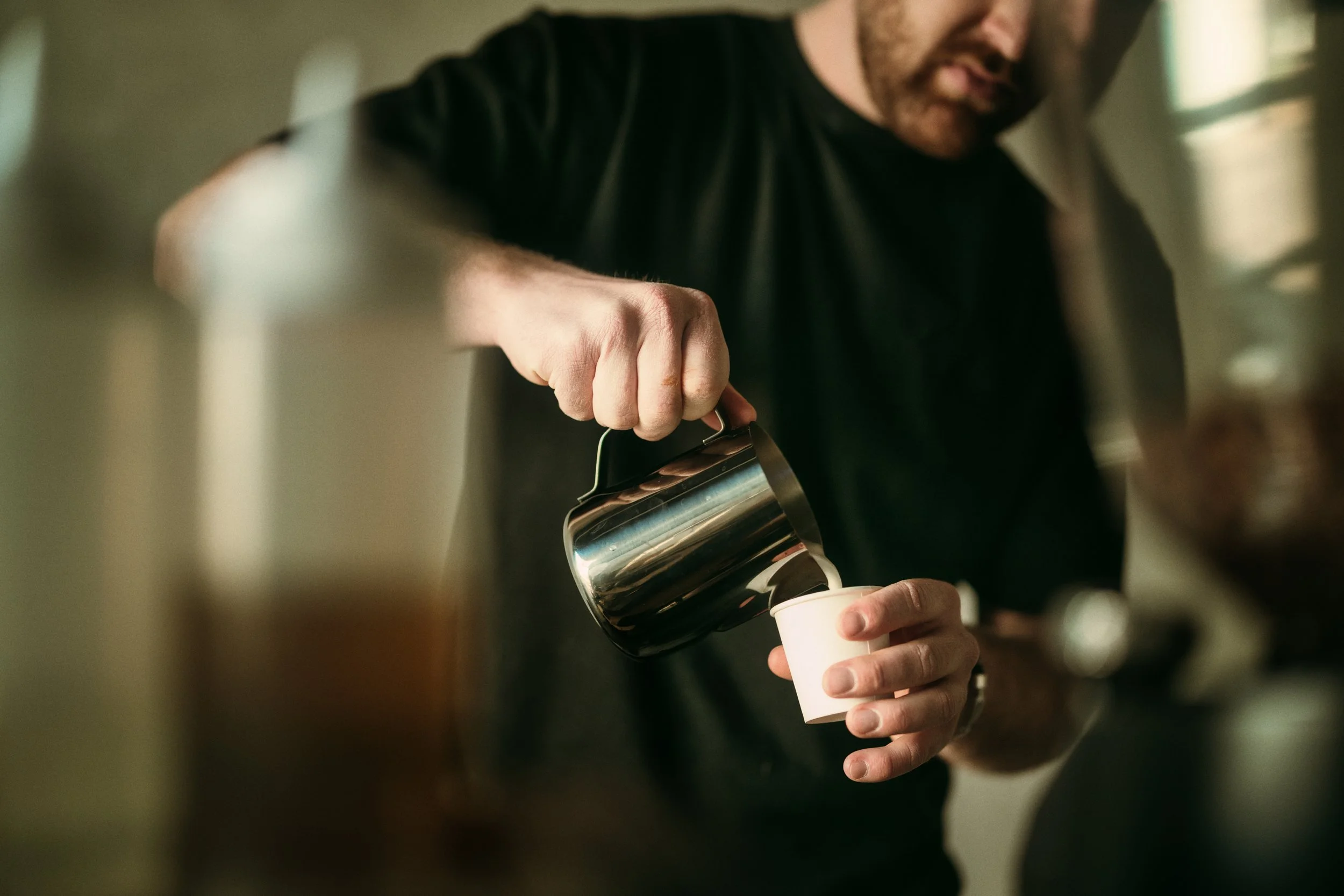 A man wearing a black shirt pouring coffee from a metal pot into a small white cup.