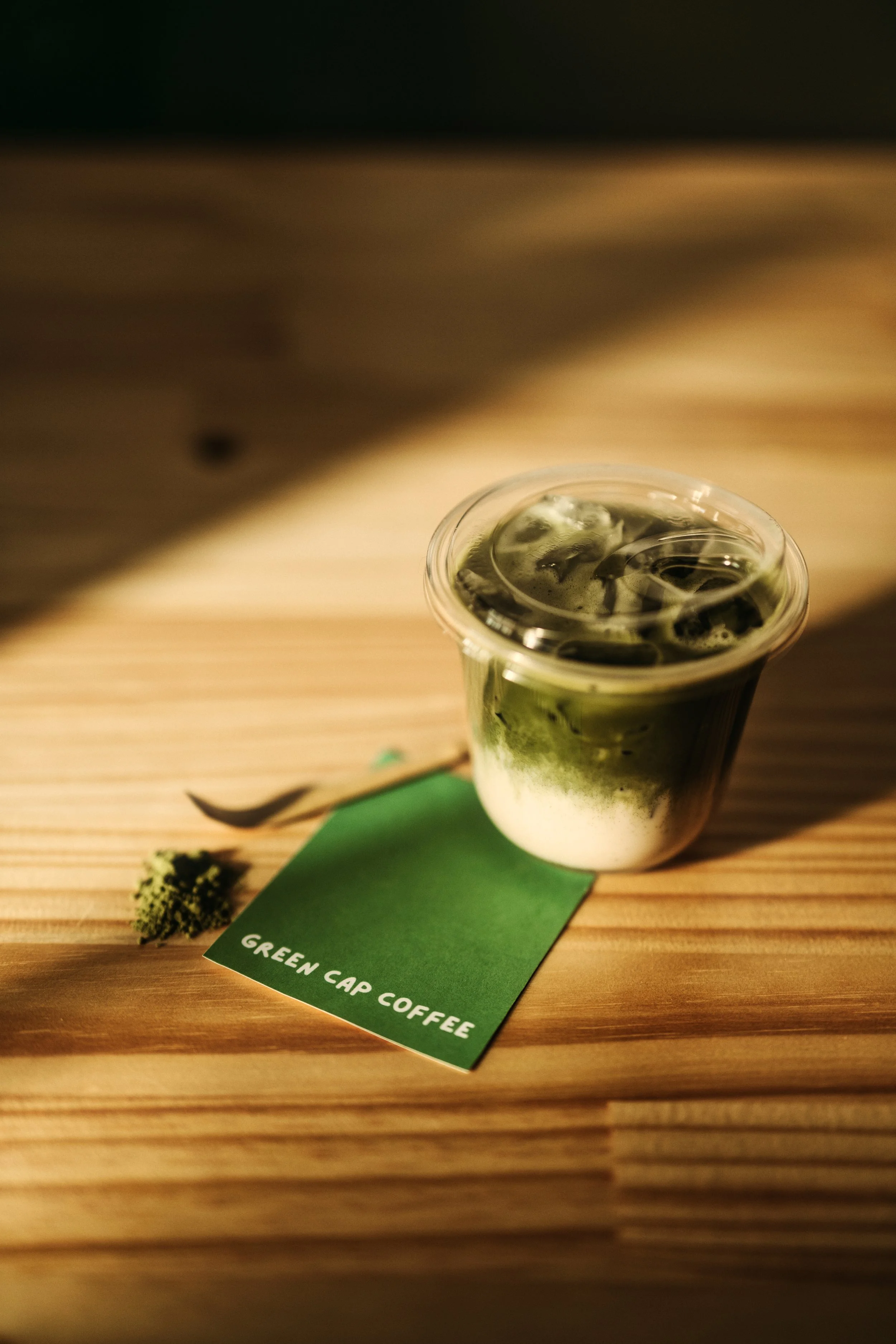 A plastic cup of green matcha latte with ice on a wooden surface, labeled with a green tag that reads 'Green Cap Coffee'.