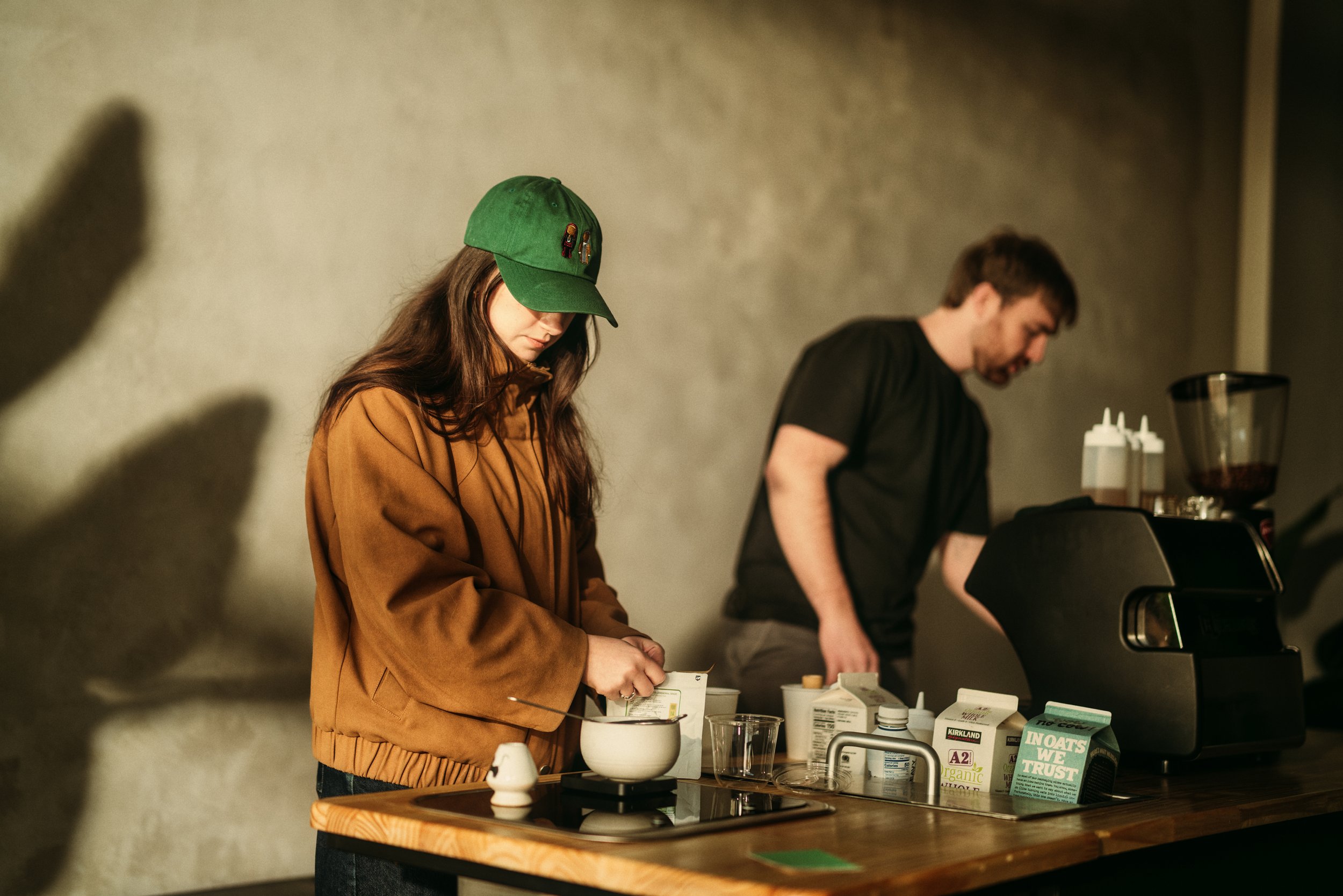 A woman wearing a green cap and brown jacket preparing a drink in a white mug at a wooden counter, with a man in a black shirt in the background operating a coffee machine.