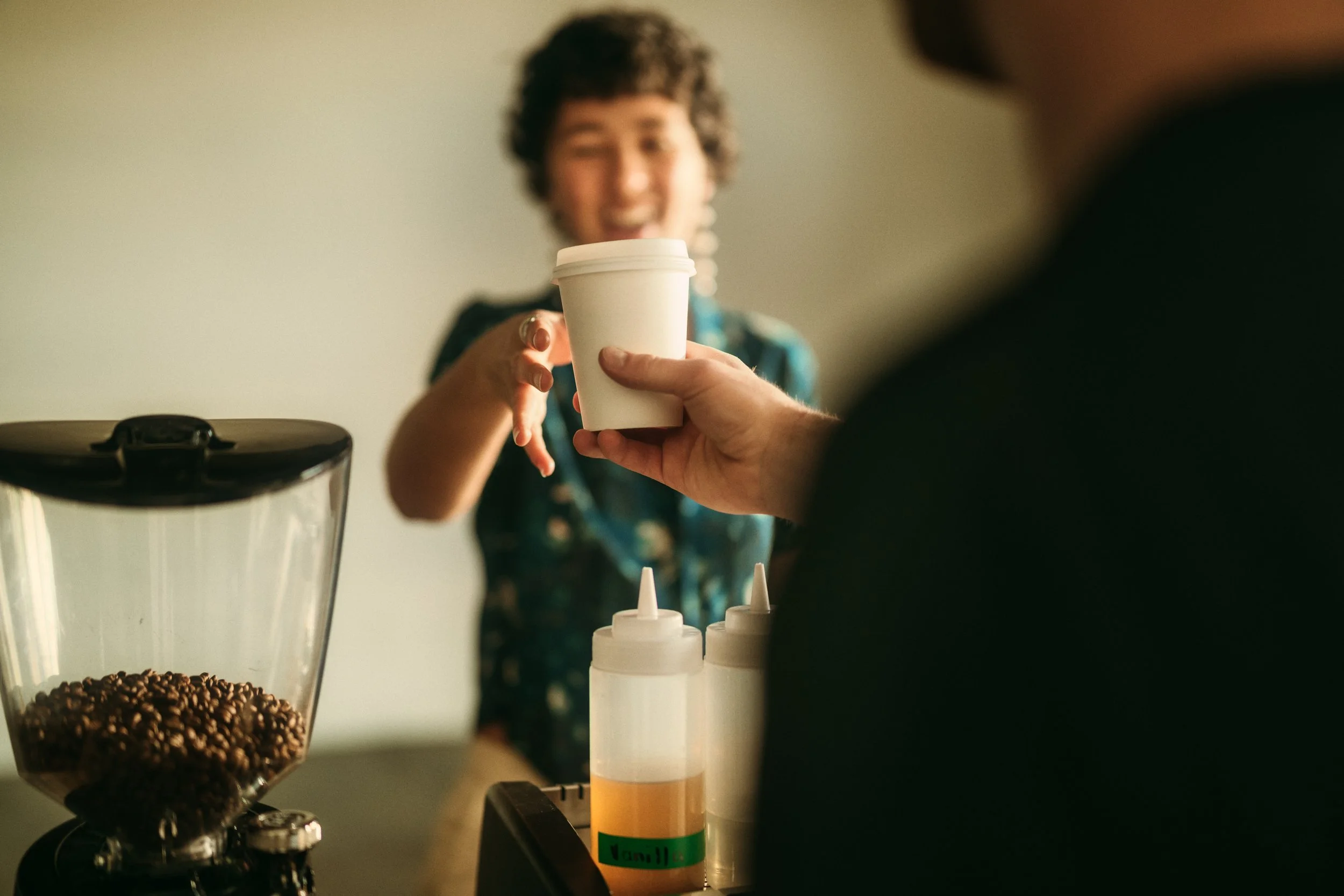 Person handing a paper coffee cup to another person at a cafe counter with coffee beans and syrup bottles nearby.