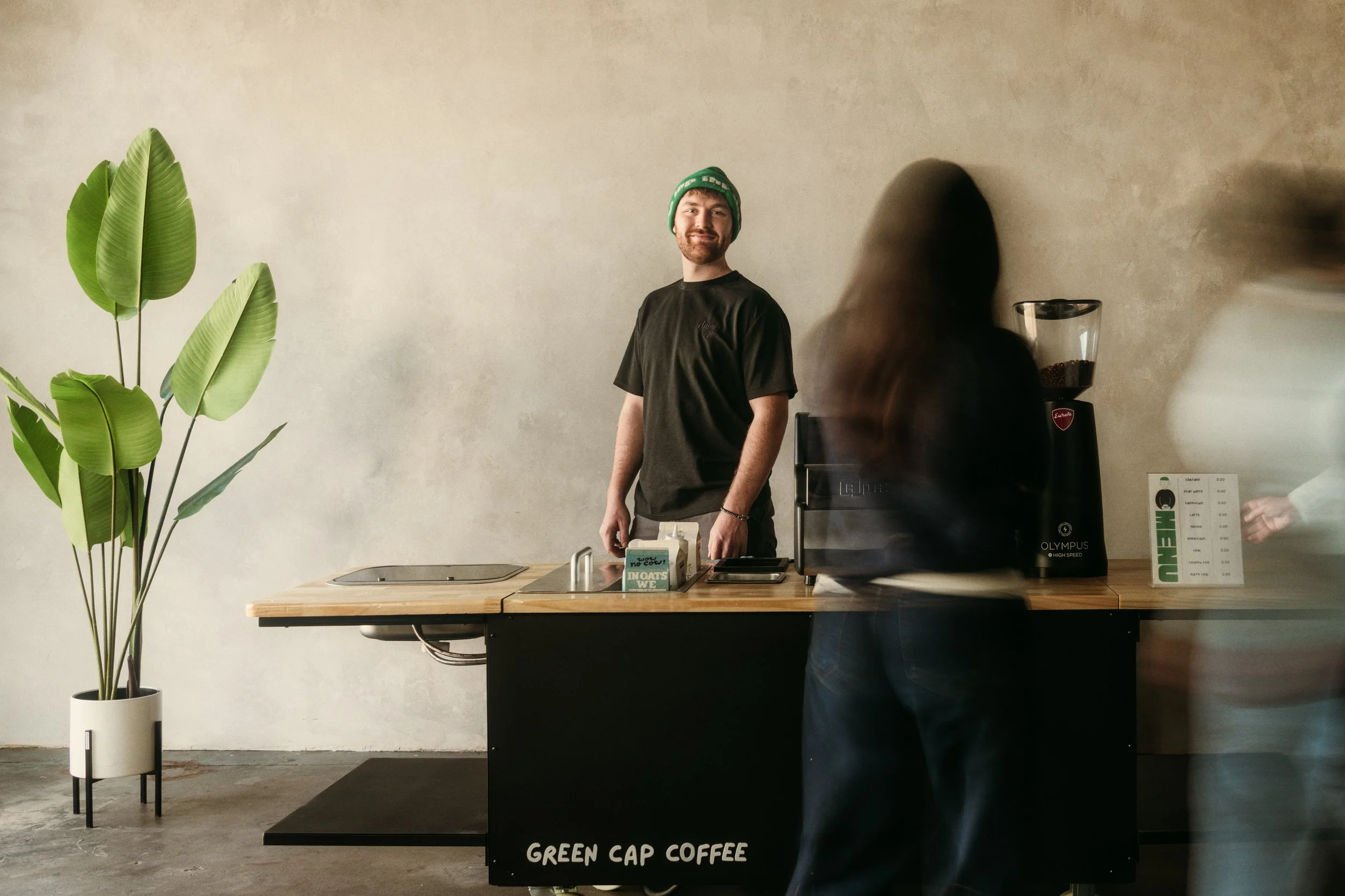 A man with a beard and wearing a green beanie, black t-shirt, and brown pants standing behind a coffee counter. There are two women in front of the counter, one with long dark hair and the other with short hair, both blurred due to movement. The coffee counter has a sign that reads "Green Cap Coffee," a coffee grinder, and a small plant on the left side. The background is a plain, beige wall.