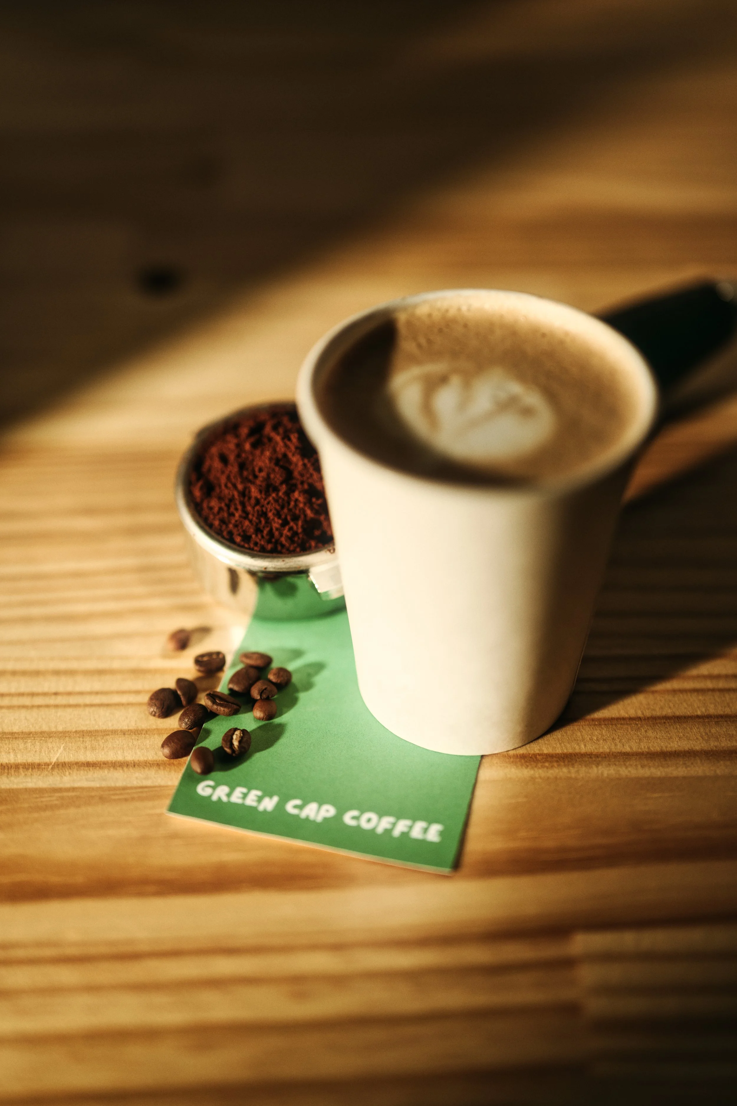 A cup of coffee with foam, a container of coffee ground, and coffee beans on a green card labeled "Green Cap Coffee" on a wooden table.