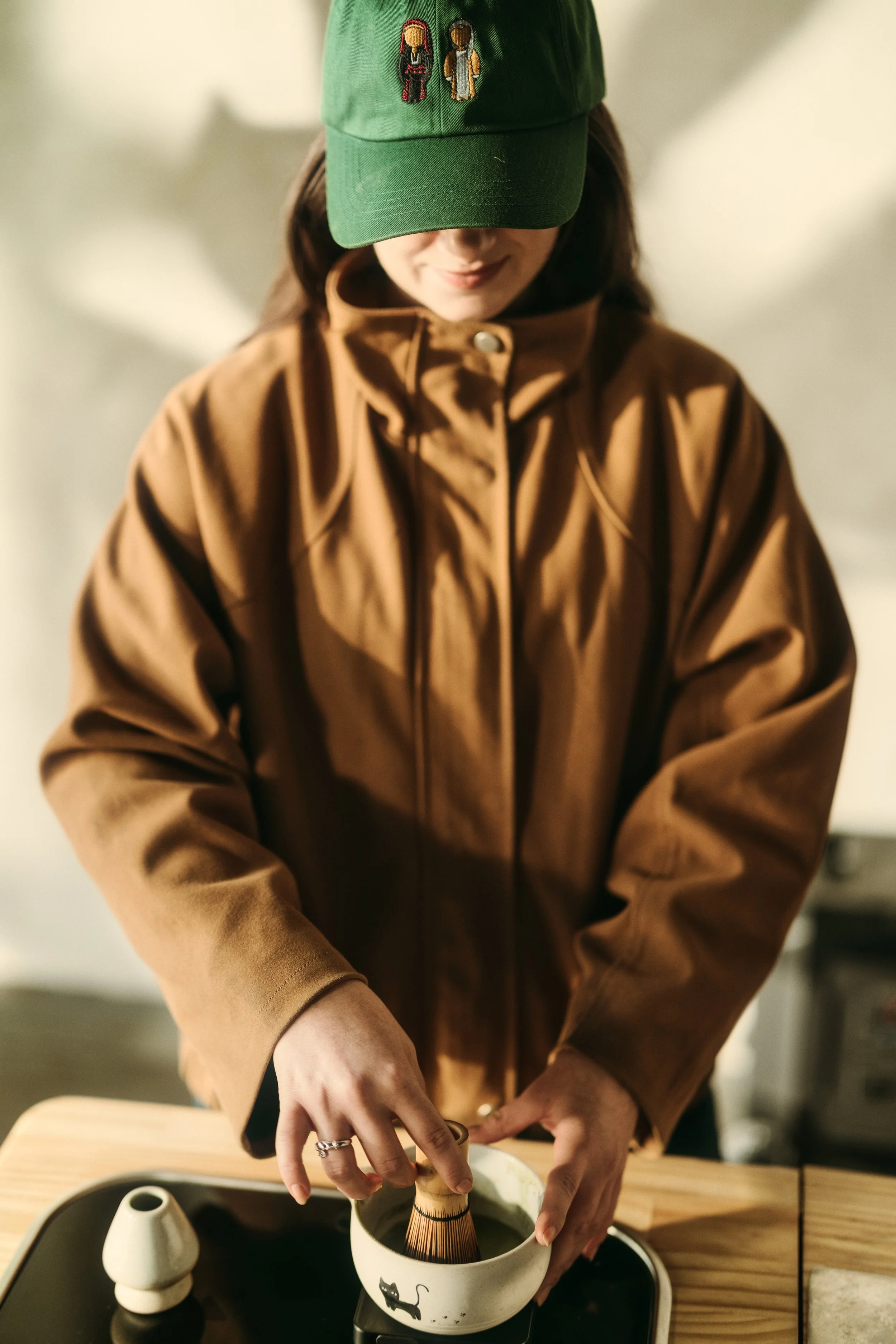 A woman wearing a green cap with embroidered characters, a brown jacket, and rings, is holding a matcha whisk and preparing to whisk matcha in a ceramic bowl with a black cat design.