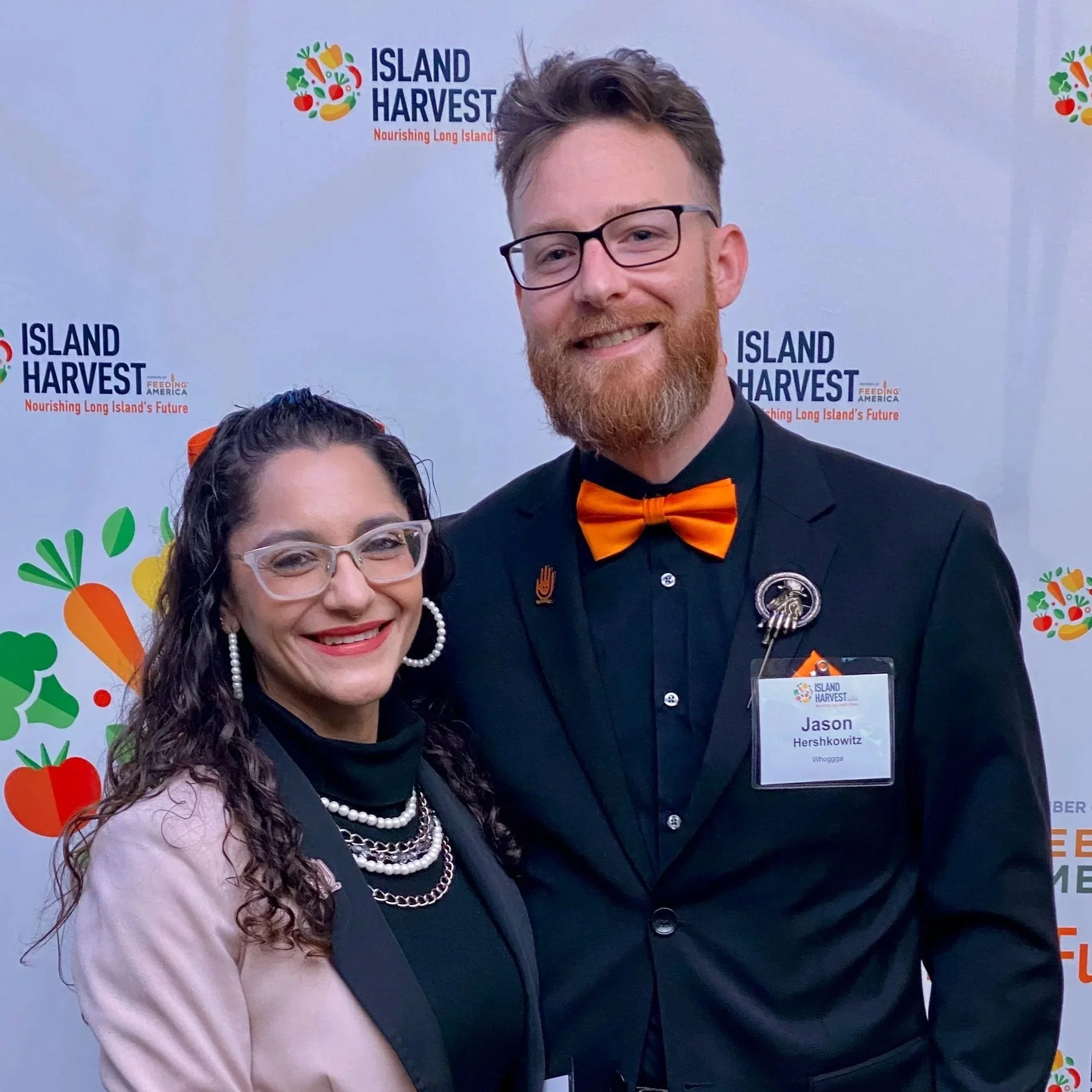 Two people smiling at an event with a background displaying "Island Harvest" logo and colorful vegetables. The woman has curly brown hair, glasses, and is wearing a light pink blazer and layered pearl necklaces. The man has a beard, glasses, and is dressed in a black suit with an orange bow tie, wearing a name tag that reads "Jason Hershkowitz".