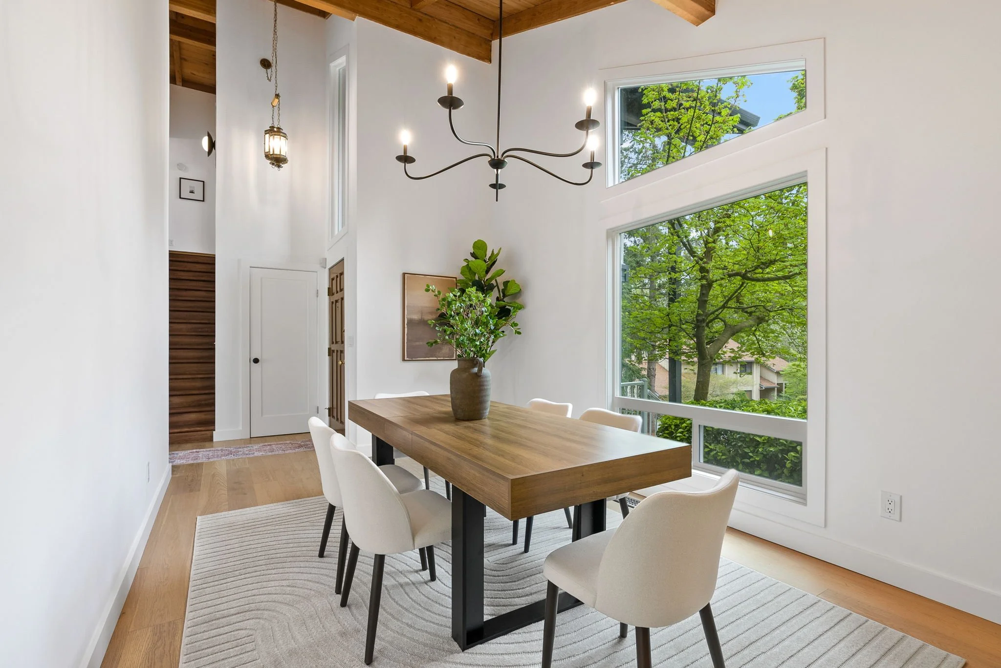 Modern dining room with white walls, large windows showing green trees outside, a wooden dining table with six white chairs, a large vase with greenery, a black chandelier, and a wooden ceiling with exposed beams.