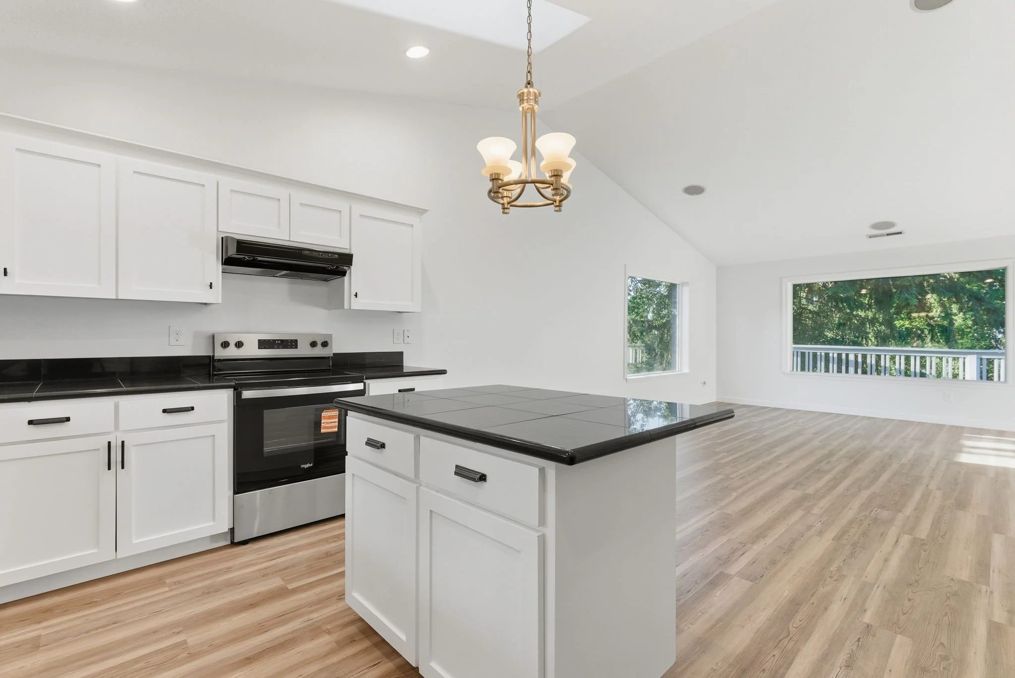 Empty kitchen with white cabinets, black countertops, black stove, and a kitchen island, leading into a bright open living space with large windows and wood flooring.