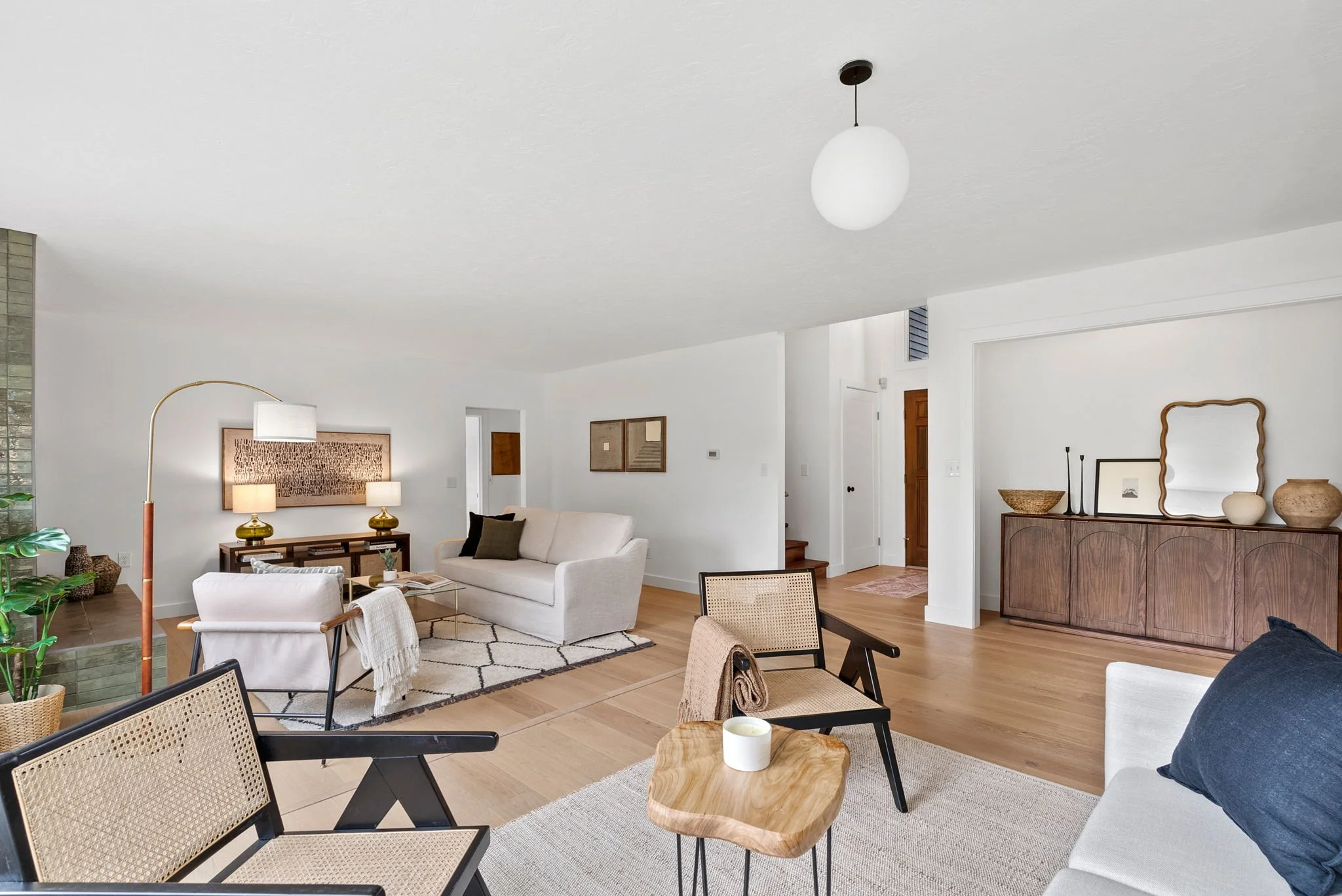 A spacious living room with modern and minimalist decor, featuring white walls, wooden floors, a sofa, armchairs, a wooden sideboard, and various decorative items.