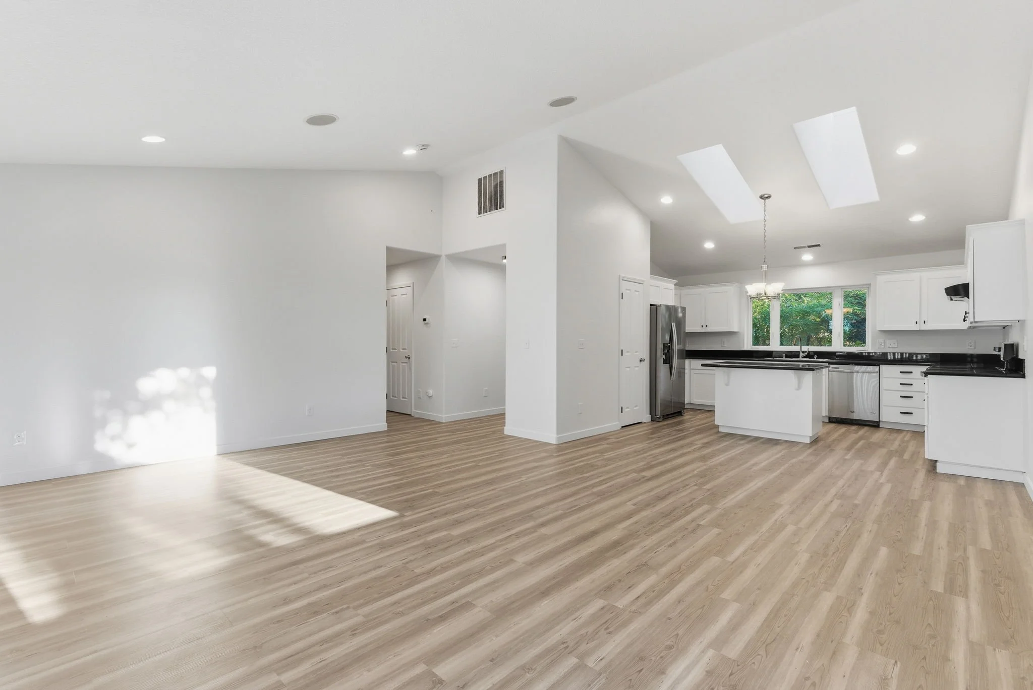 Spacious, bright open-concept kitchen and living area with white walls, white cabinetry, black countertops, stainless steel refrigerator, hardwood floors, and large window with greenery outside.