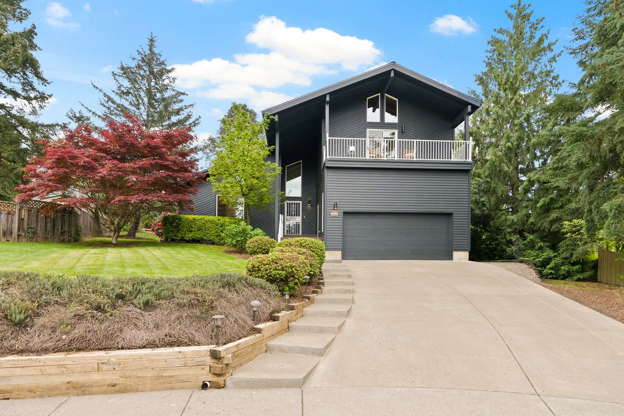Modern two-story house painted dark gray with a garage, front steps, green lawn, trees, and landscaping on a sunny day.