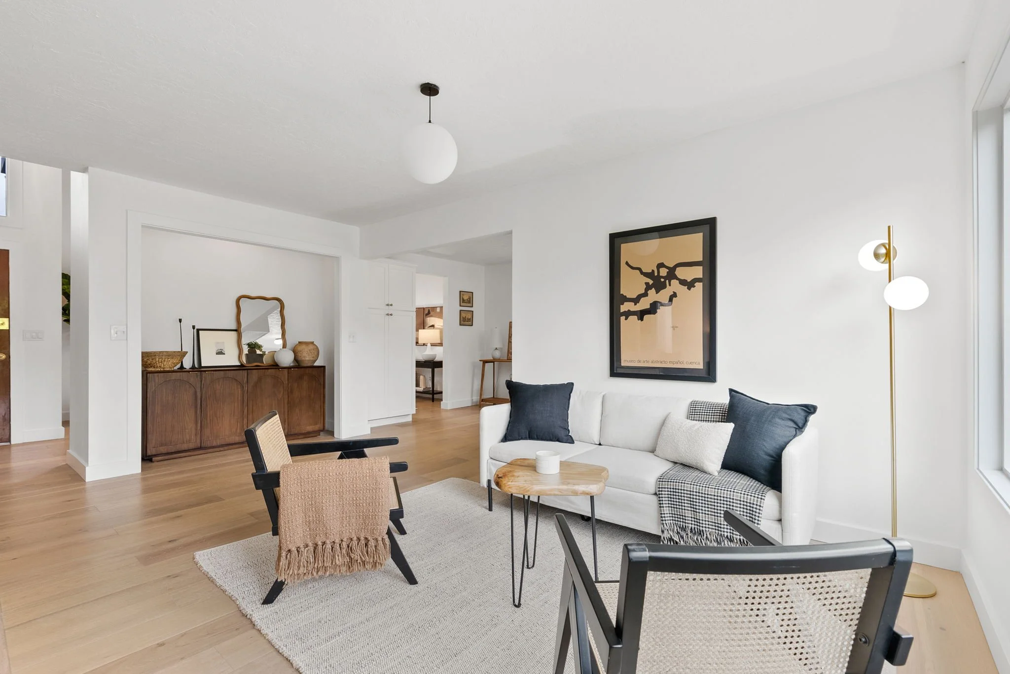 Modern living room with white walls, hardwood flooring, a white sofa with black and white pillows, a wooden side table, black chairs with woven seating, a floor lamp with two white globes, a framed abstract art piece on the wall, and a textured beige