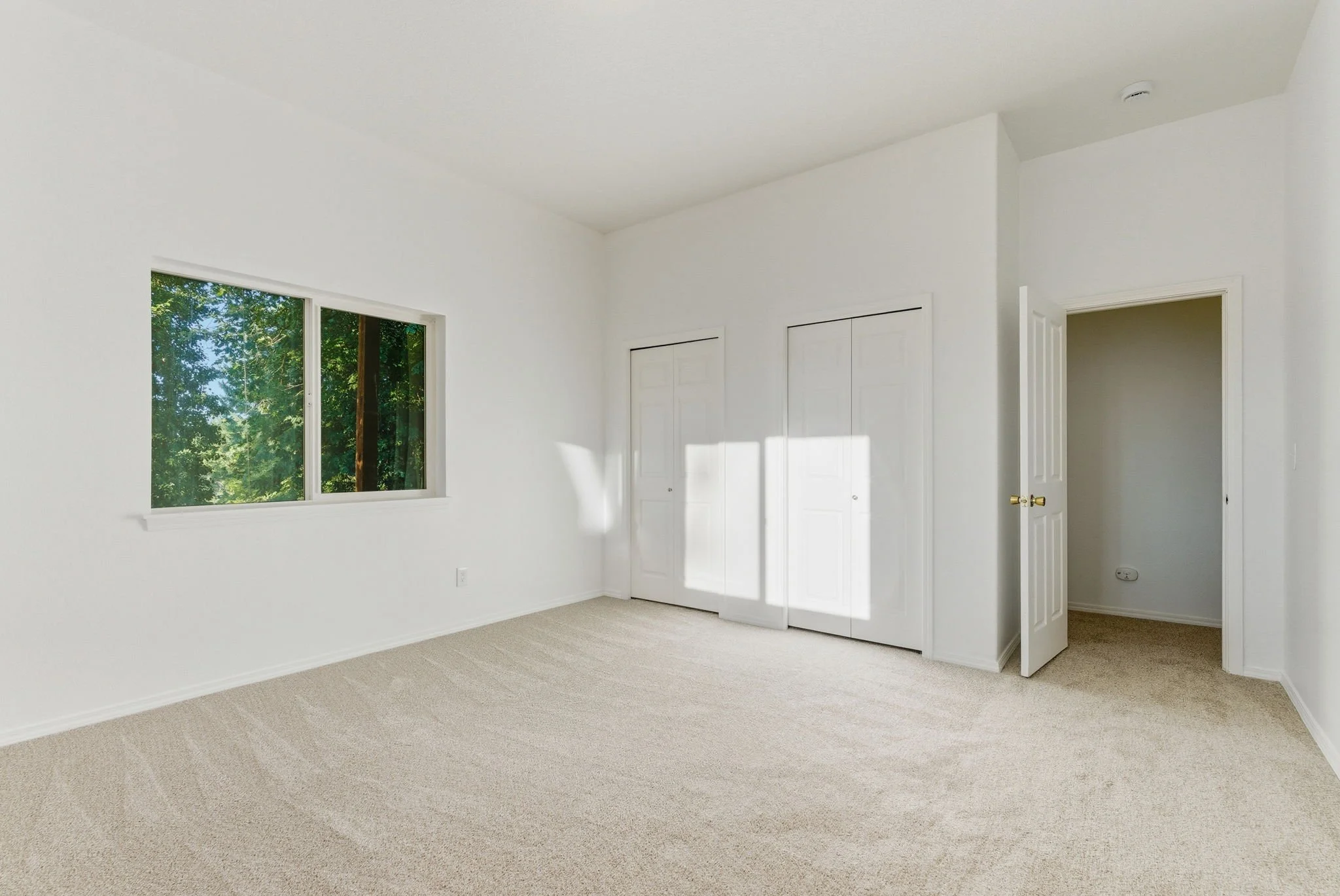 Empty room with a window, closet doors, and an open door leading to a small space, all with white walls and beige carpet.