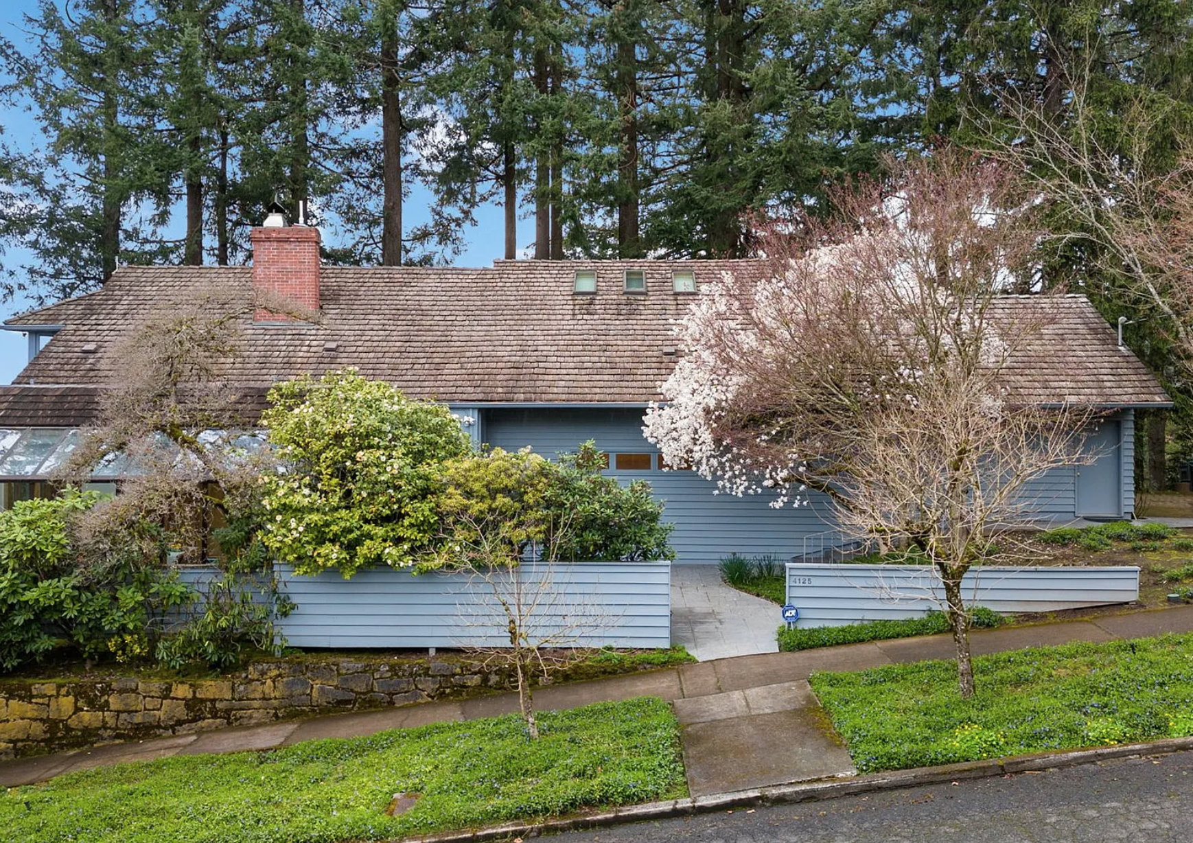 A house with a brown tiled roof, brick chimney, and light blue siding, surrounded by trees and shrubs, with a concrete walkway in front.