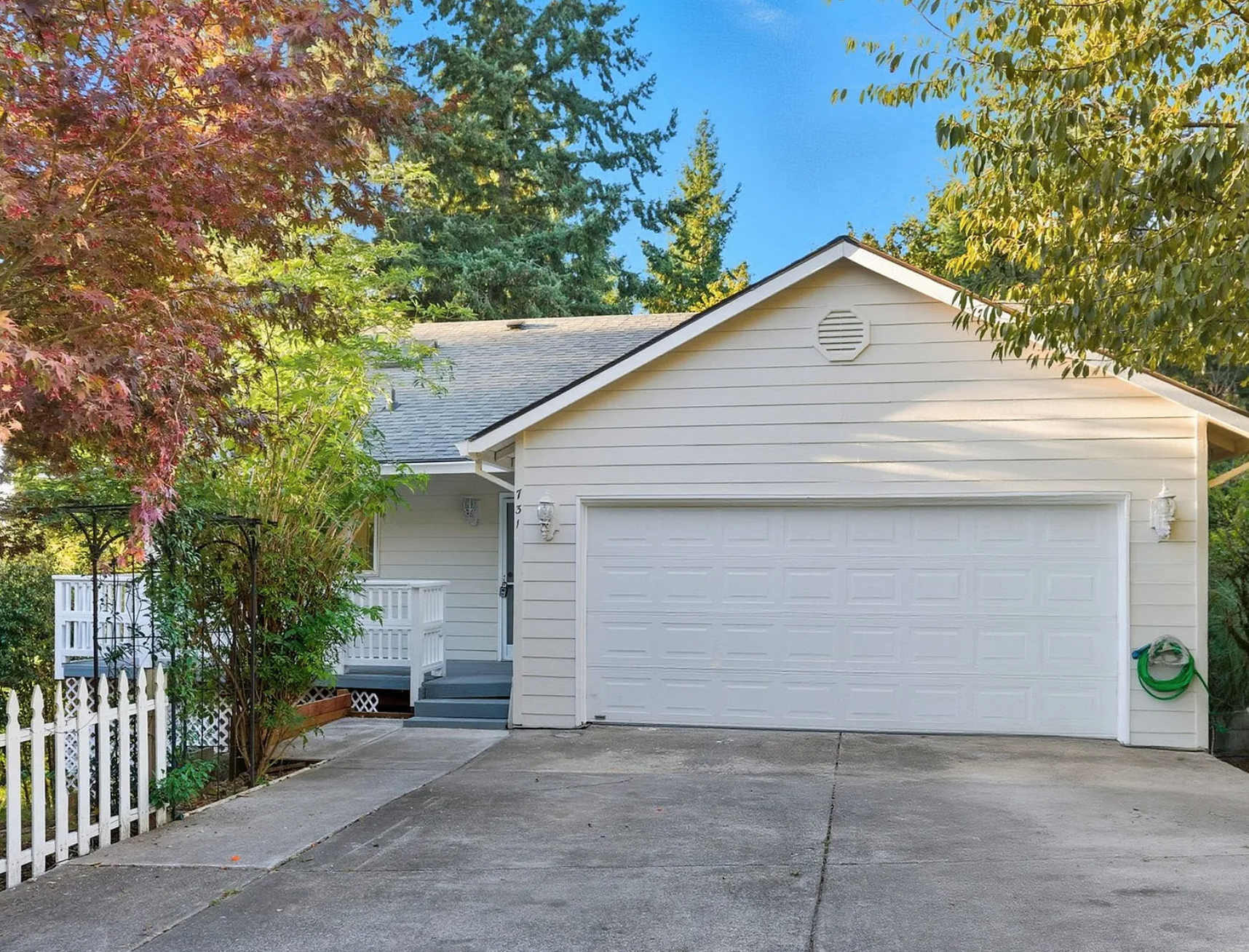 White house with a garage door, small front porch, surrounded by trees and plants, with a concrete driveway.