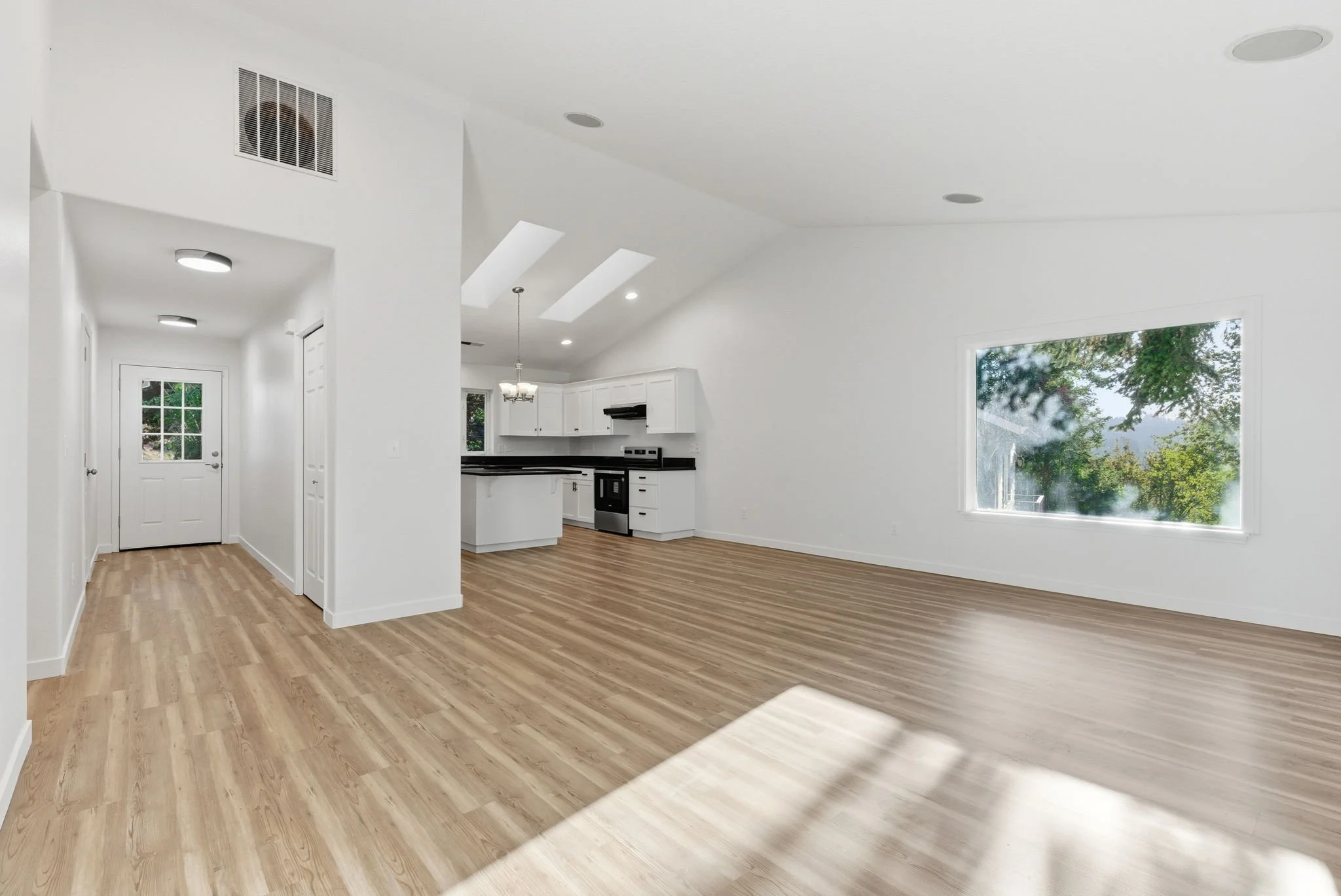 Empty living room with large window, white walls, light wood flooring, and open kitchen area with white cabinets and black countertops.