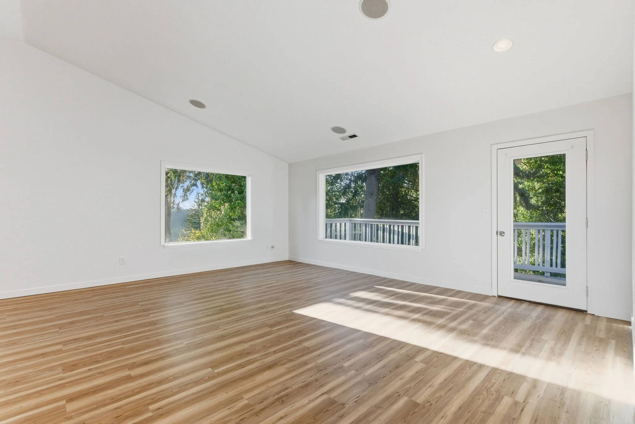Empty room with white walls, wooden floors, large windows, and a glass door leading to a balcony with trees outside.
