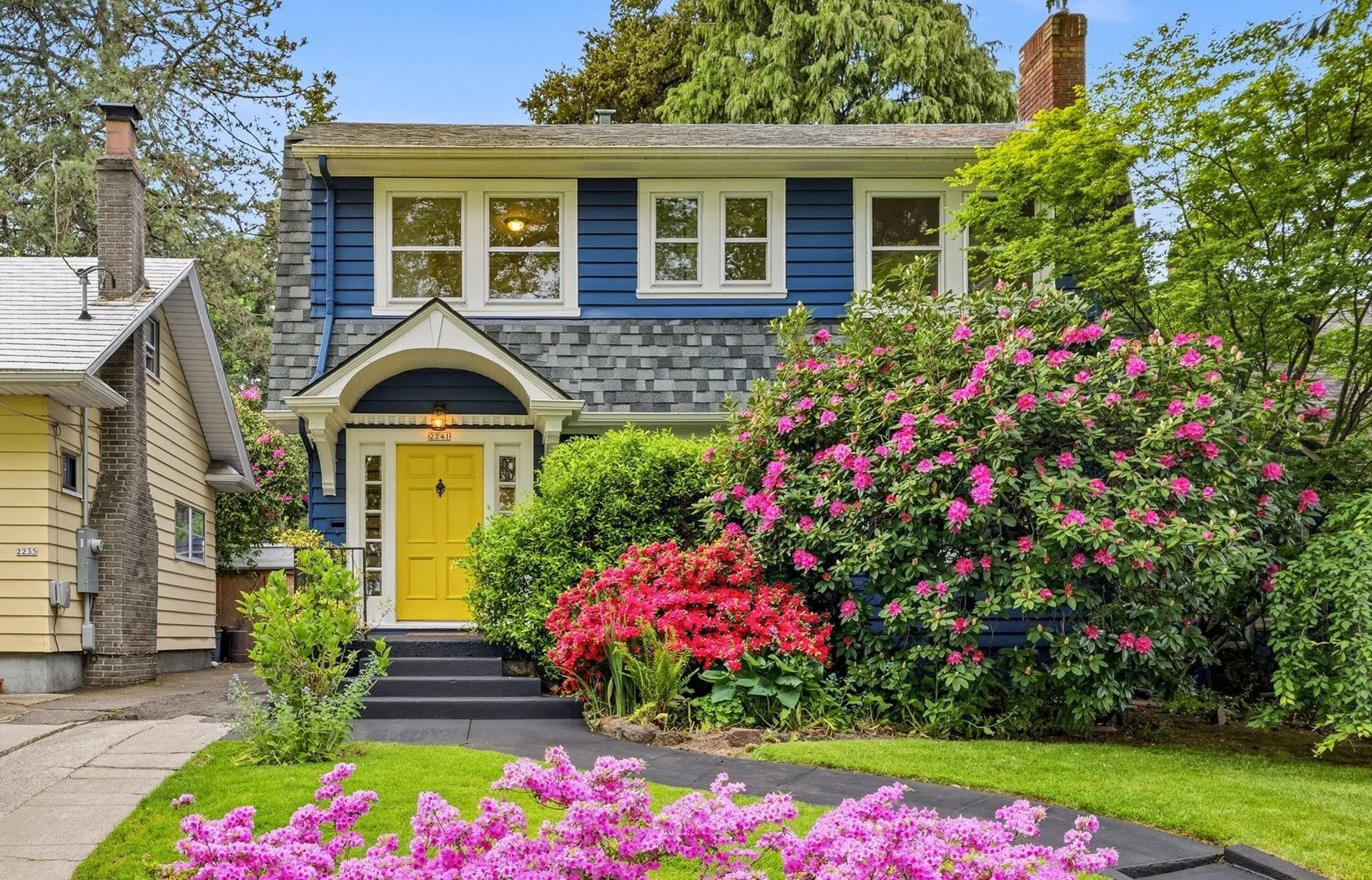 A two-story house with blue siding and a yellow front door, surrounded by a lush garden with pink and purple flowering bushes, green grass, and trees, under a clear blue sky.