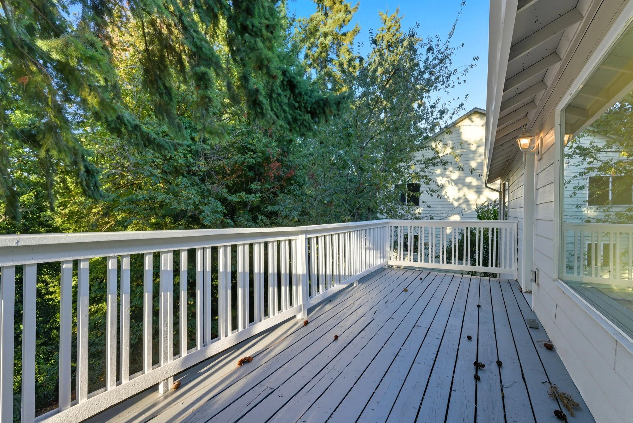 A wooden balcony with white railing attached to a house, with a view of trees and neighboring houses in the background, and sunlight shining on the house wall.