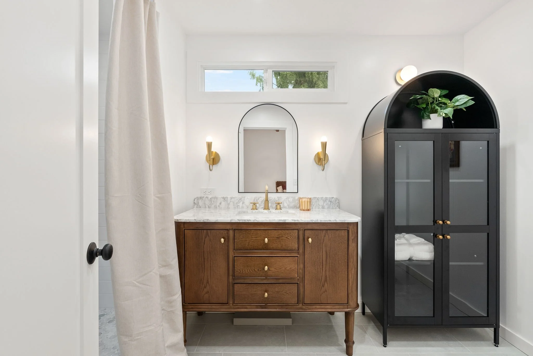 Bathroom with white walls, a marble-top wooden vanity with gold fixtures, a mirror, two wall sconces, a small window, and a black cabinet with a potted plant inside.