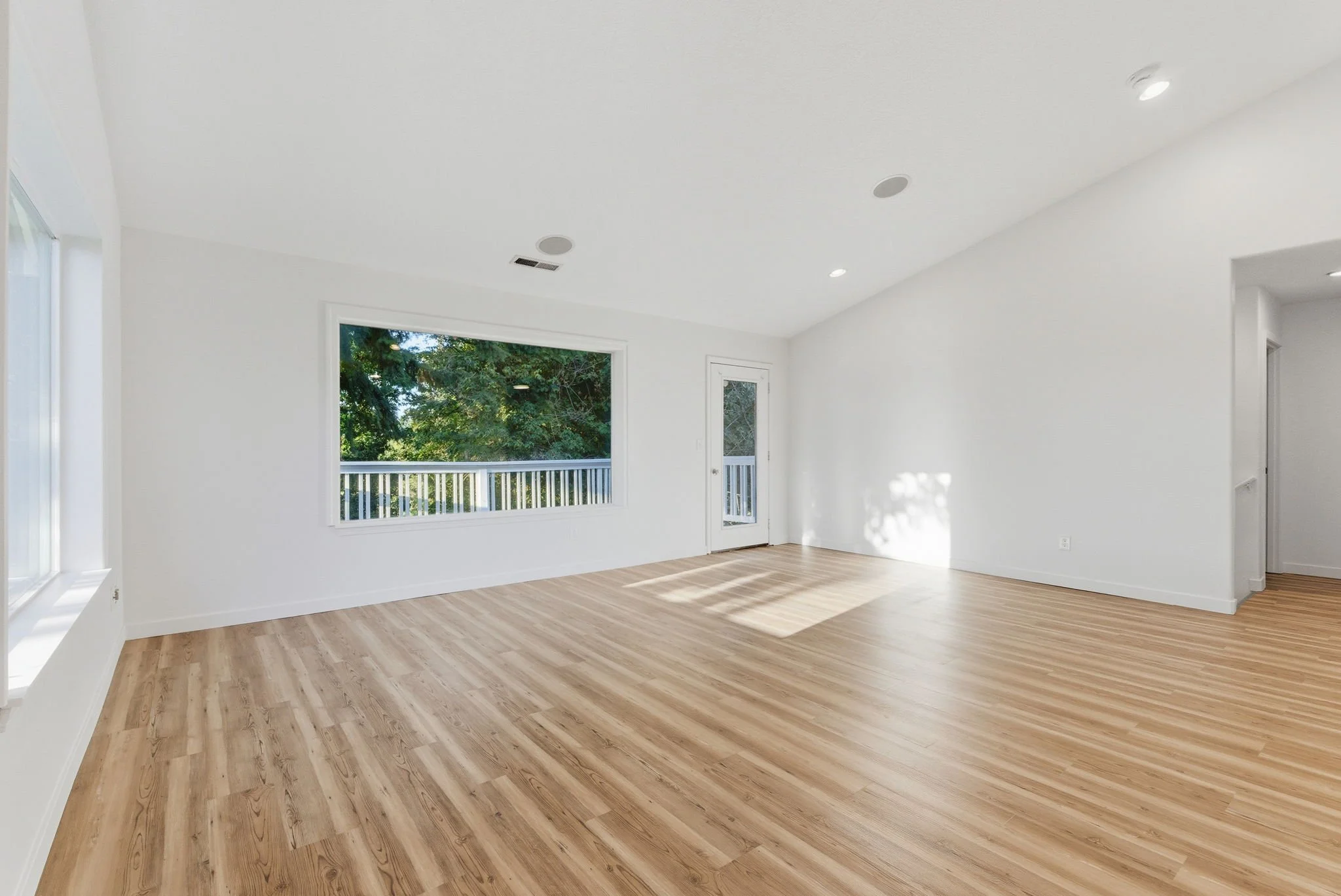 Empty living room with white walls, light wood flooring, large window, and glass door leading to outside deck.
