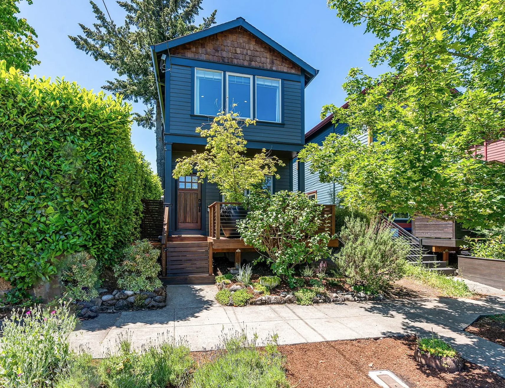 A two-story blue house with wooden accents, large front windows, and a small porch with steps, surrounded by lush green trees and shrubs, with a sidewalk in the foreground.
