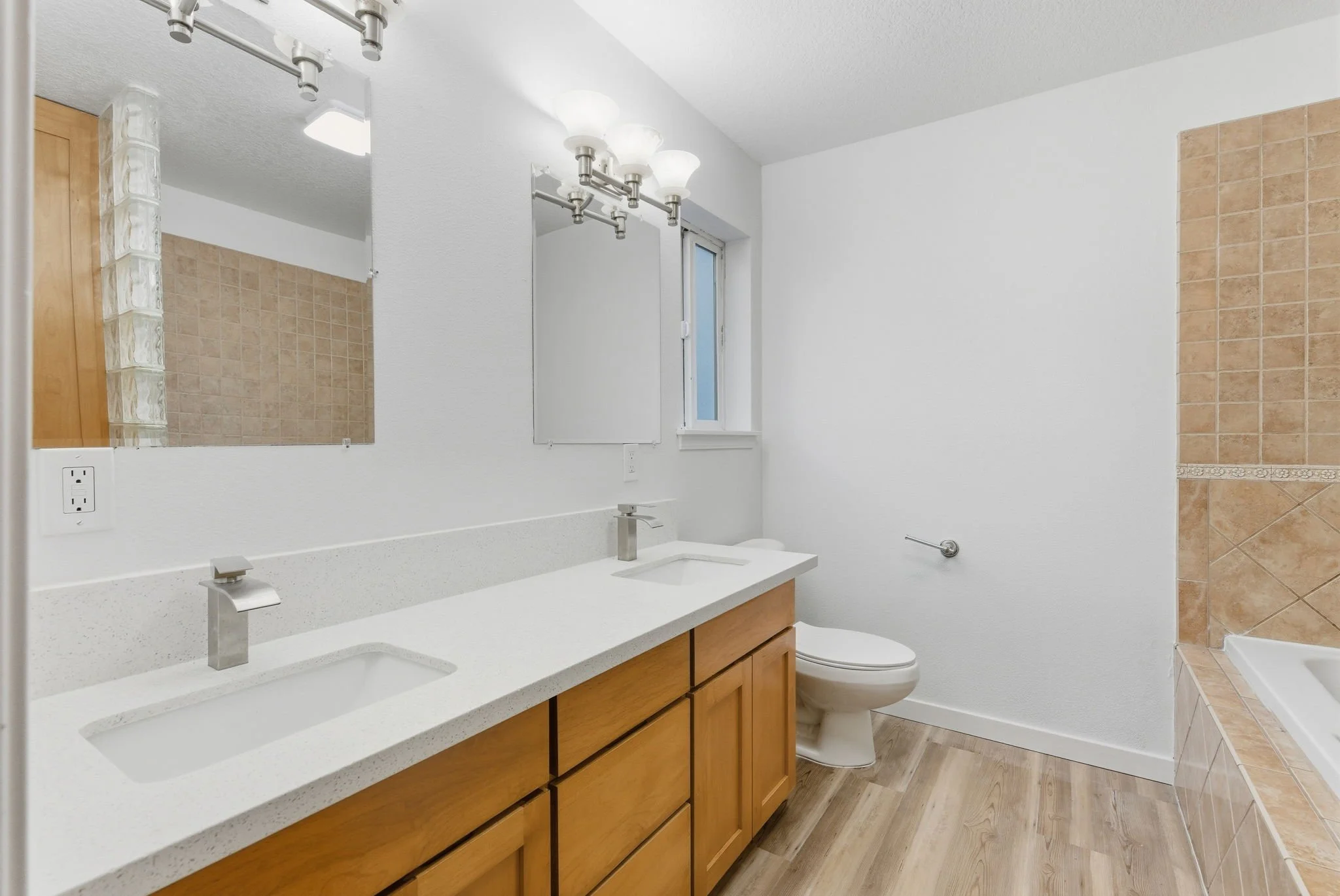 A clean bathroom with a double vanity sink, a large mirror, a small window, a toilet, and a bathtub with beige tile surround.
