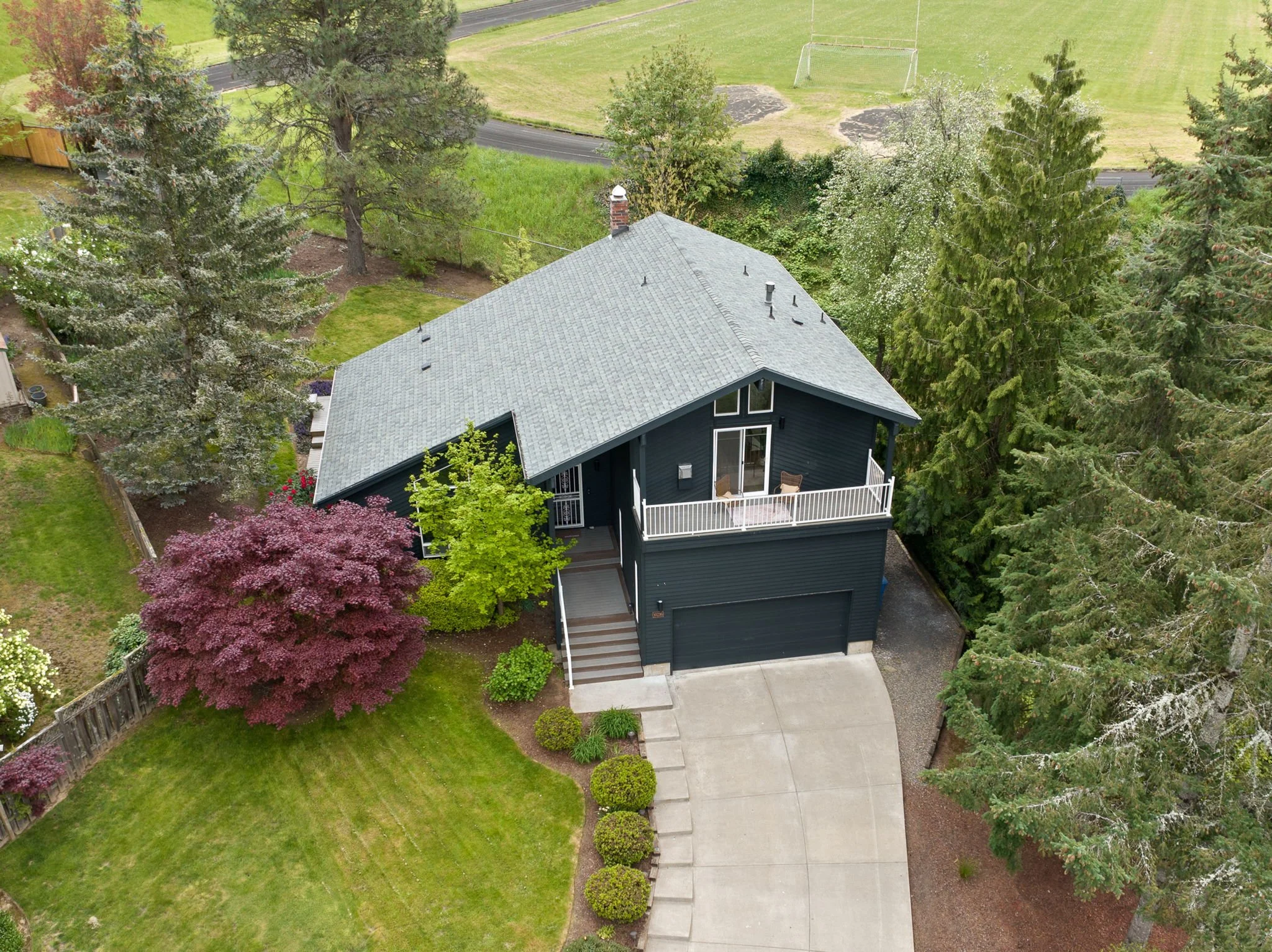 Aerial view of a dark blue two-story house with a gray roof, front porch, and attached garage, surrounded by trees and a well-maintained lawn.