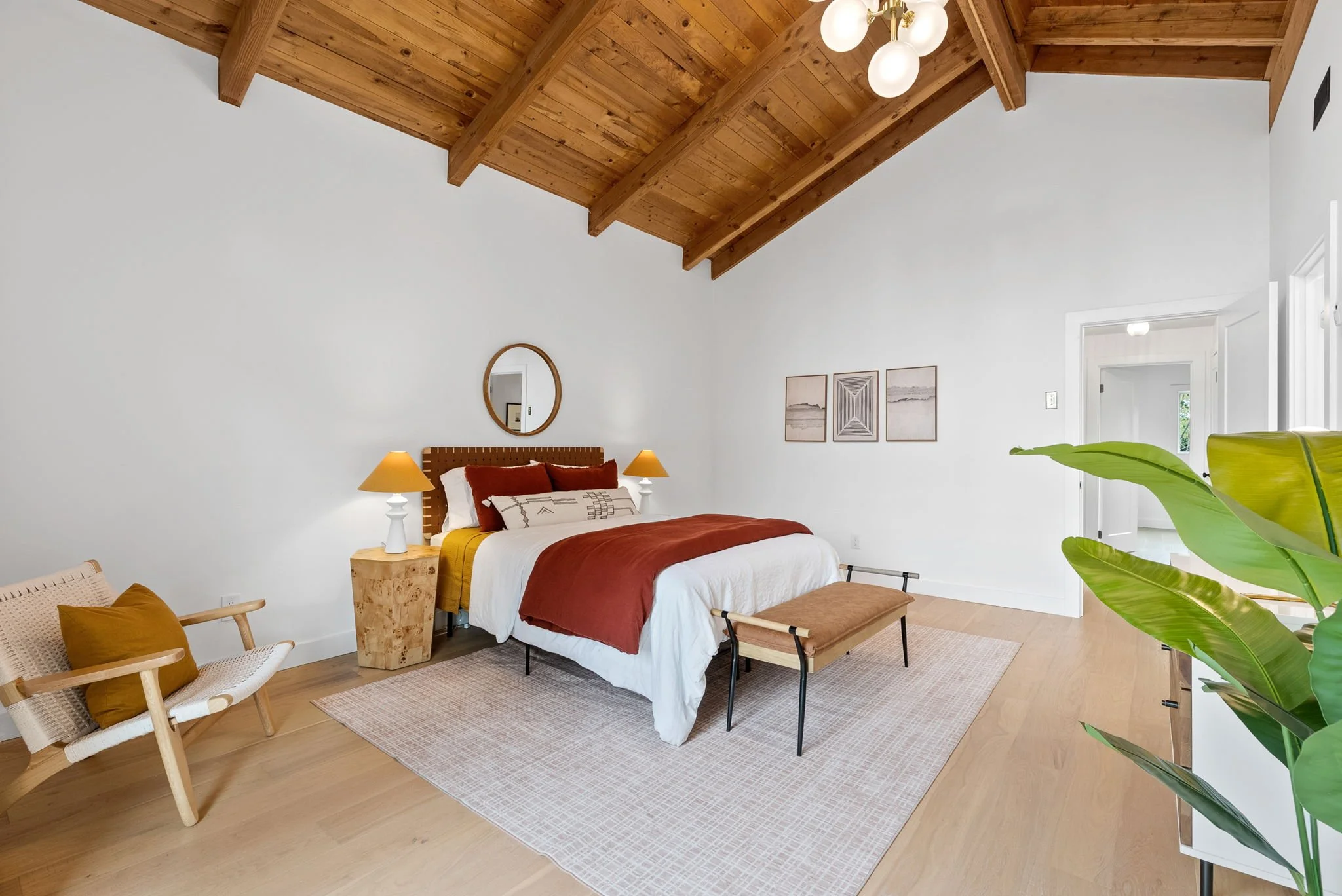 A bedroom with a high wooden ceiling, white walls, and hardwood floors. It has a bed with a wooden headboard, white bedding, rust-colored pillows, and a mustard and rust-colored throw. There are two small bedside lamps on wooden nightstands. On the r