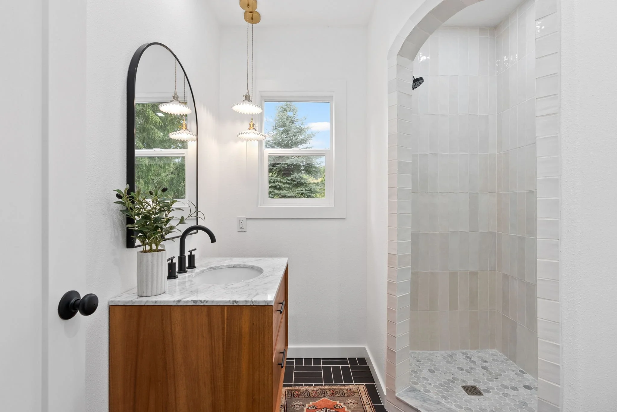 Modern bathroom with a wooden vanity, white marble countertop, potted plant, black fixtures, large mirror, pendant lighting, window with view of trees, and walk-in shower with tiled walls and pebble floor.