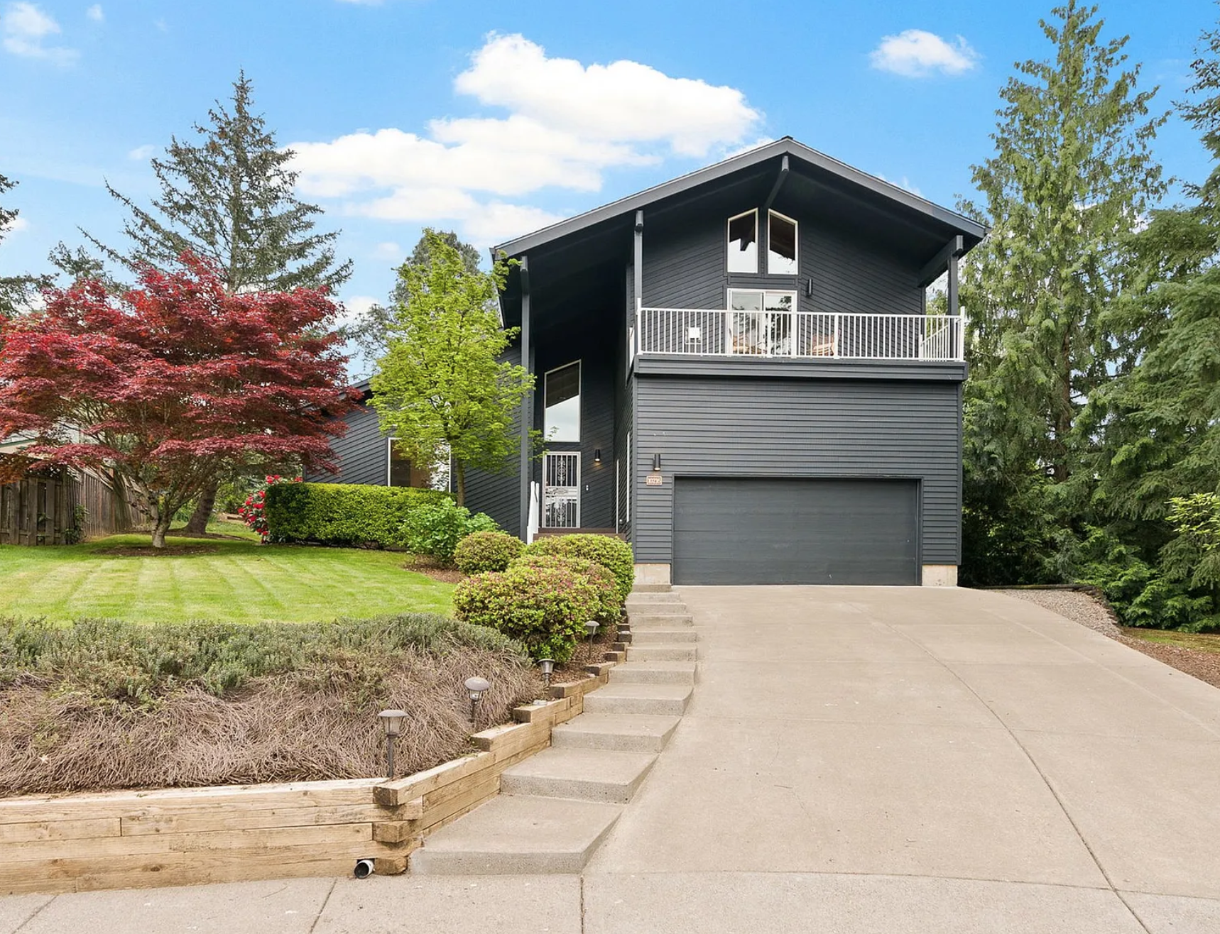 A modern black two-story house with a garage and a balcony, surrounded by a well-maintained lawn, shrubs, and trees under a blue sky.