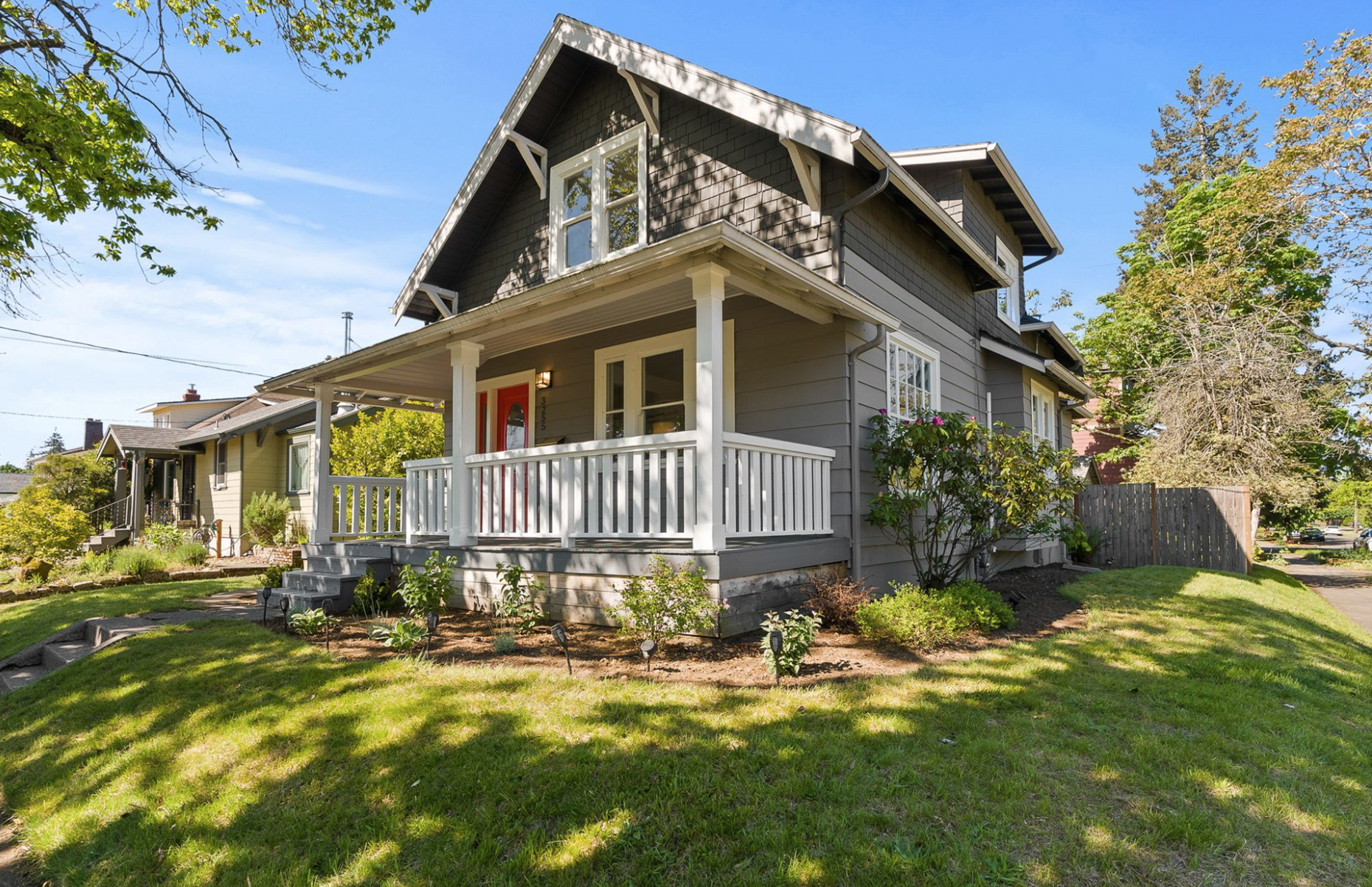 A two-story grey house with a front porch, white railing, and steps leading up to the front door, surrounded by a landscaped lawn with bushes and trees on a sunny day.