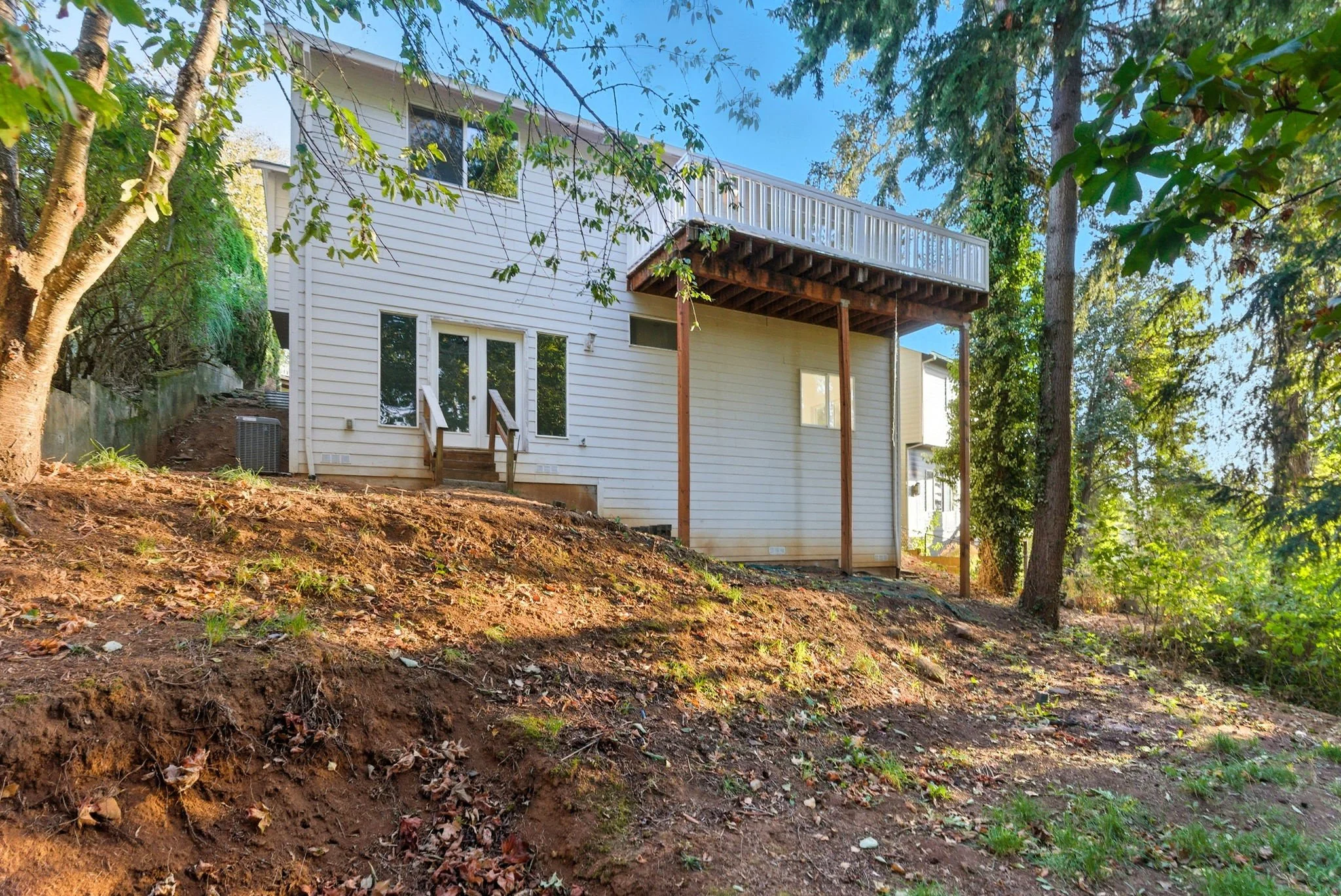 Rear view of a white two-story house with a deck on the second floor supported by wooden posts, situated on a sloped yard with trees and exposed soil.