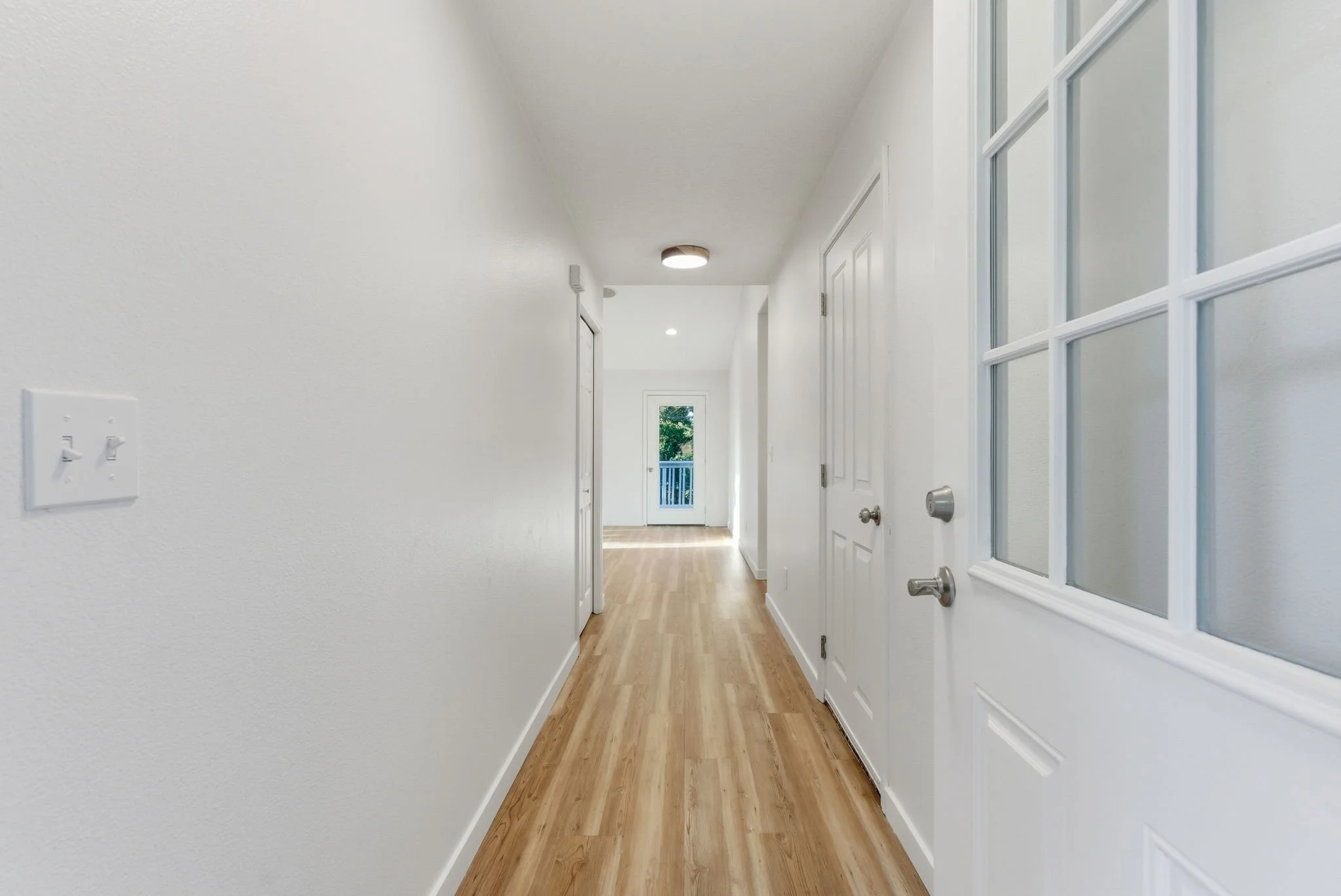 Empty hallway with white walls, wooden flooring, and multiple doors, with a door at the end leading to a balcony outside.