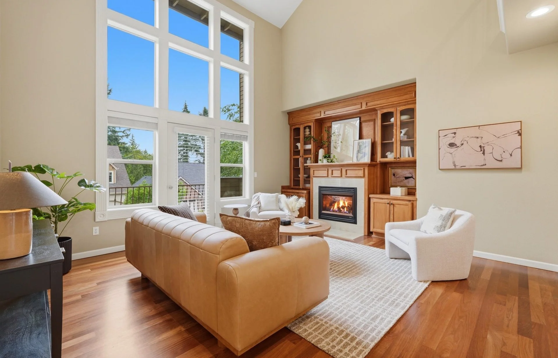 Living room with vaulted ceiling, tall two-story windows, hardwood floors, a tan leather sofa, a gas fireplace with a built-in wood entertainment center, and a treed neighborhood view.