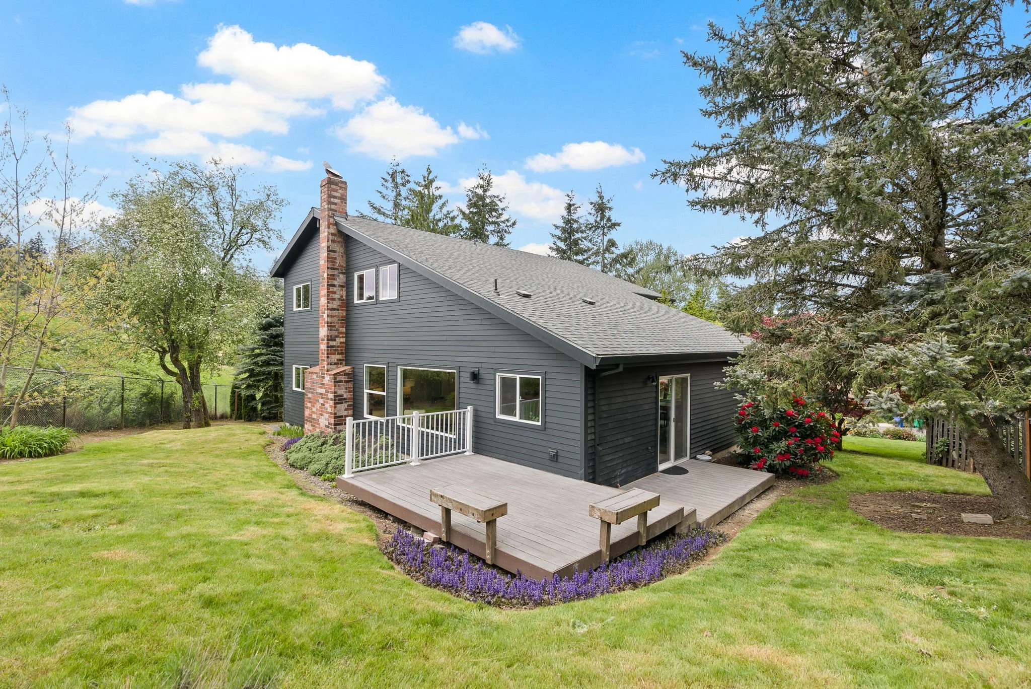 A two-story house with dark gray siding and a brick chimney in a green backyard with trees, a lawn, a wooden deck, purple flowers, and a sliding glass door, under a blue sky with clouds.