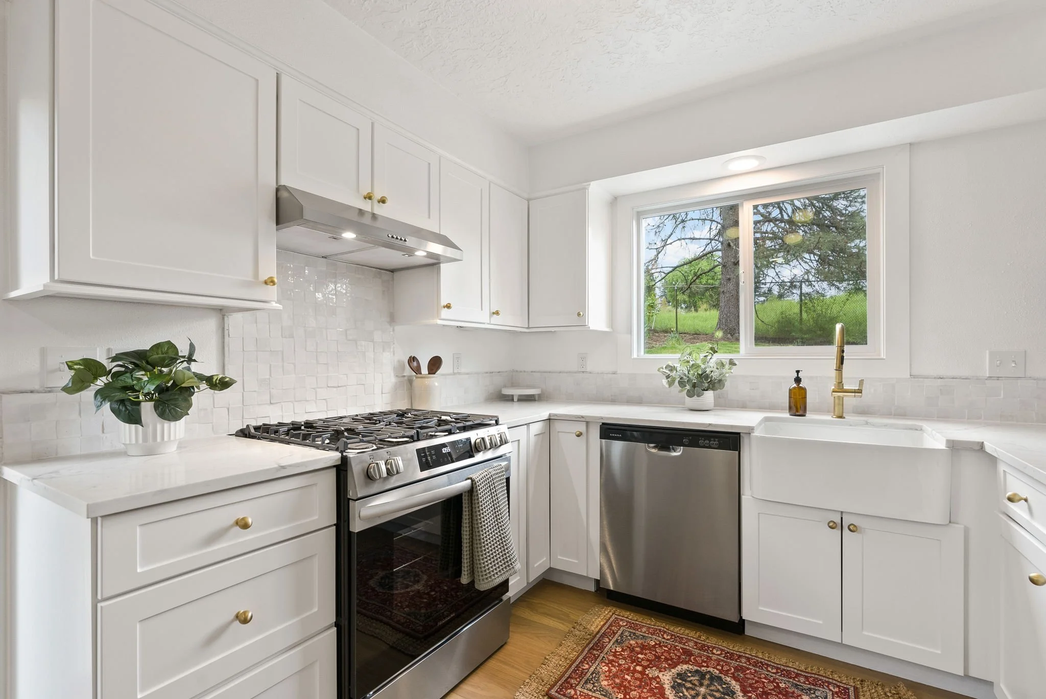 Kitchen with white cabinets, a window overlooking a green yard, a stainless steel dishwasher, a stove, and a sink with a gold faucet.