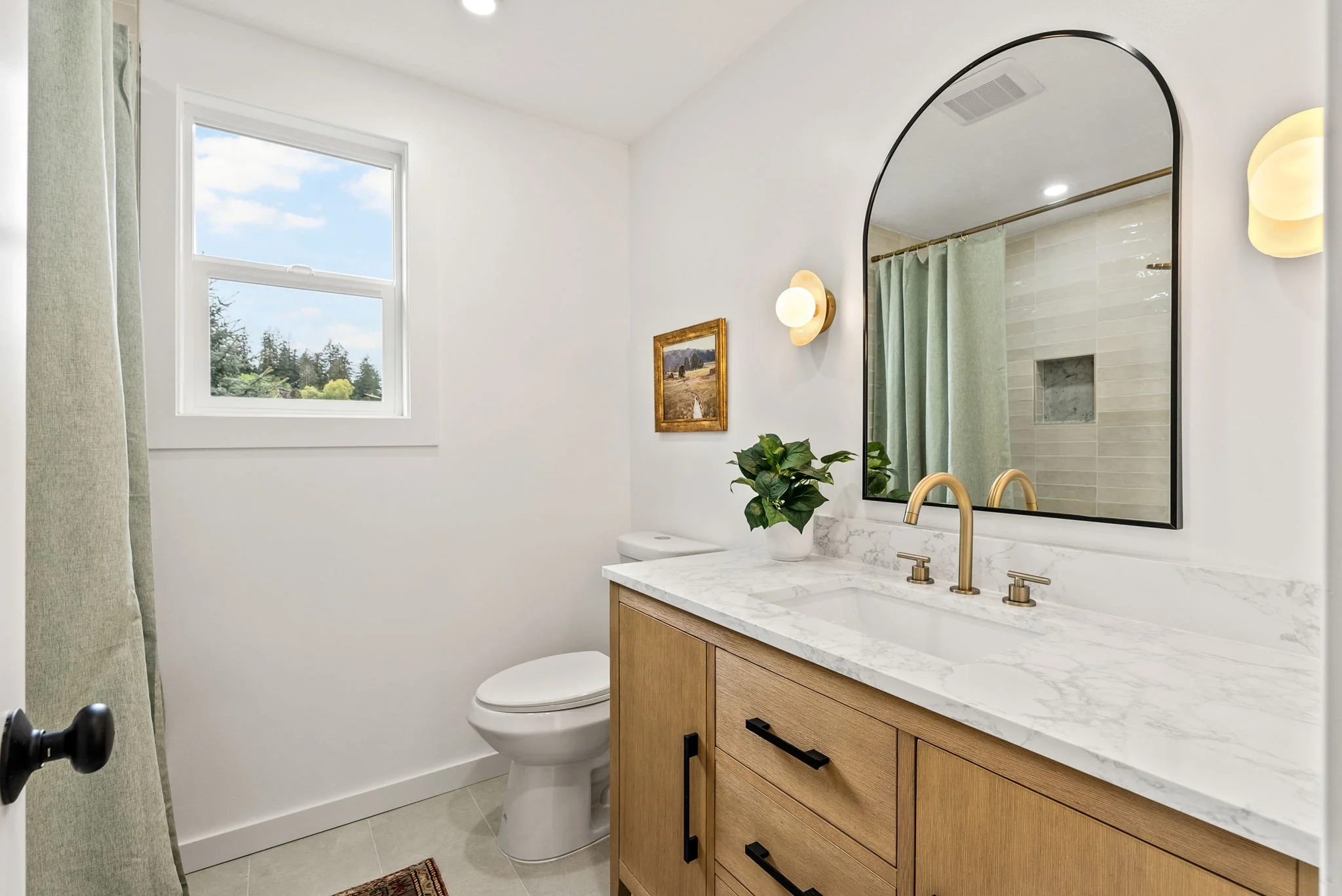 Modern bathroom with a wooden vanity, marble countertop, gold fixtures, large mirror, white walls, tile flooring, window showing sky and trees, wall-mounted lights, potted plant, shower with green curtain, and framed picture.